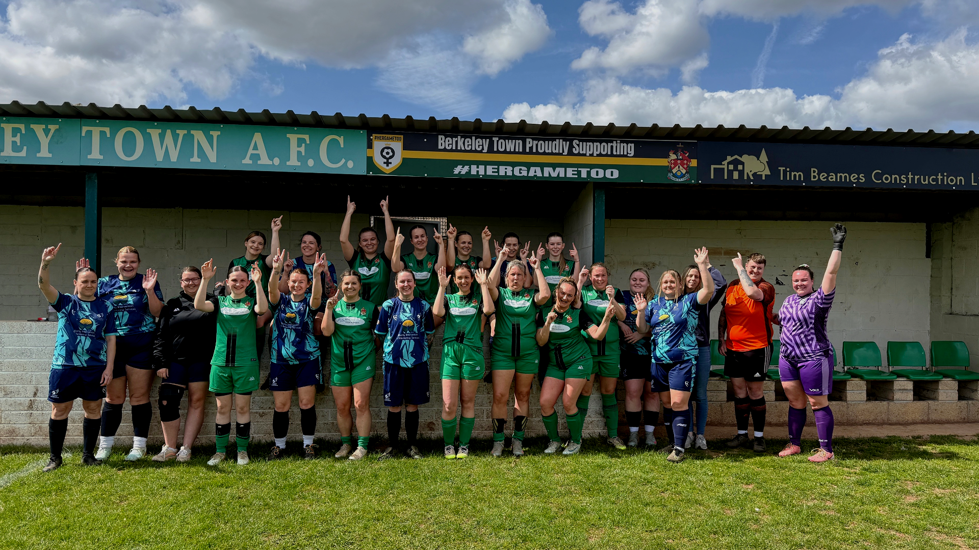 Group of women's soccer team in green, blue, and purple uniforms standing on the field, smiling and raising their hands, with a sign that reads Berkeley Town A.F.C. in the background.