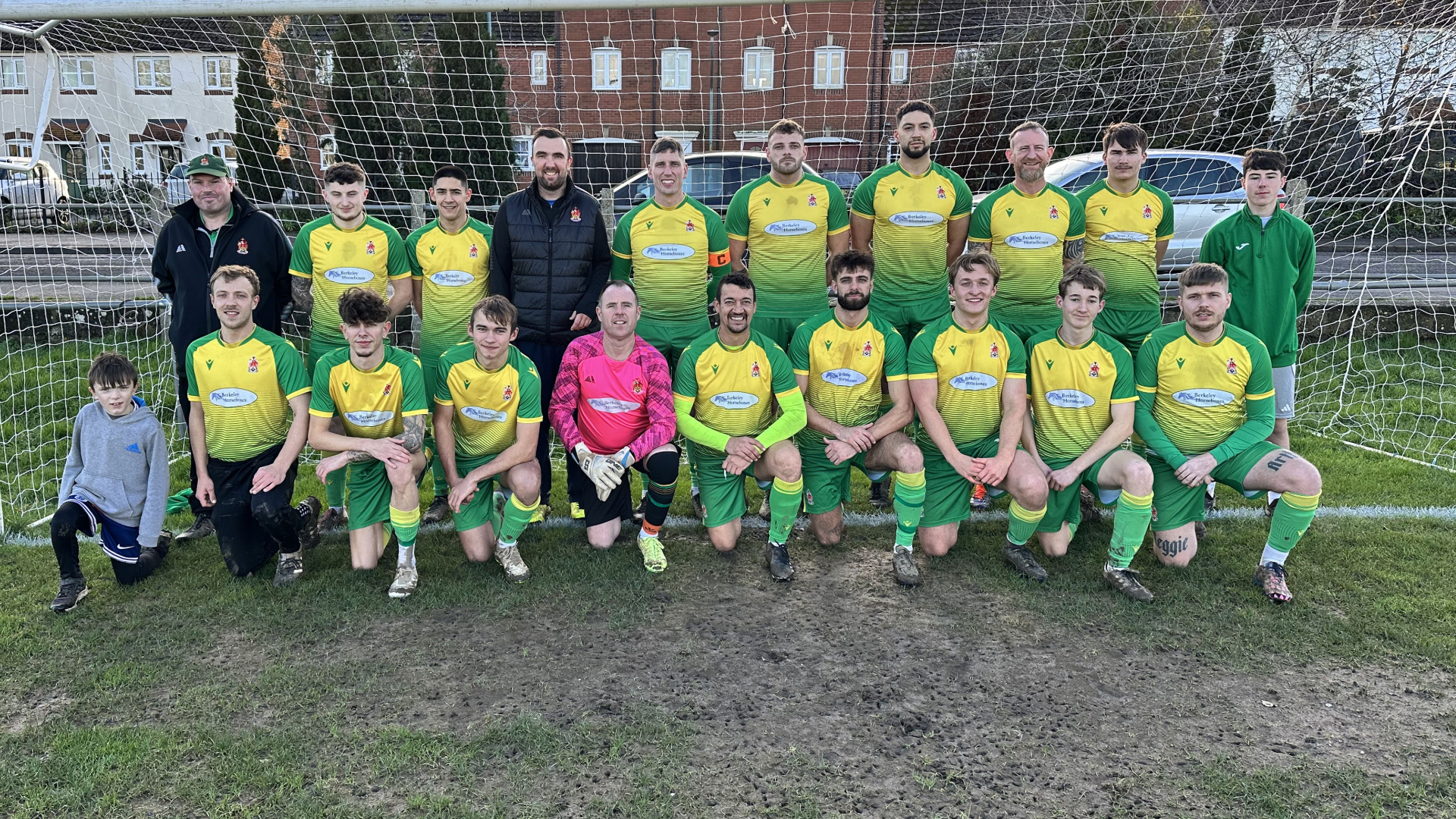 A youth soccer team pose for a group photo in front of a goal net on a grass field. The team members are dressed in yellow and green jerseys, with a few players kneeling in front and others standing behind. There are two adult coaches and a young boy in casual clothing on the left side. The background includes houses and parked cars.