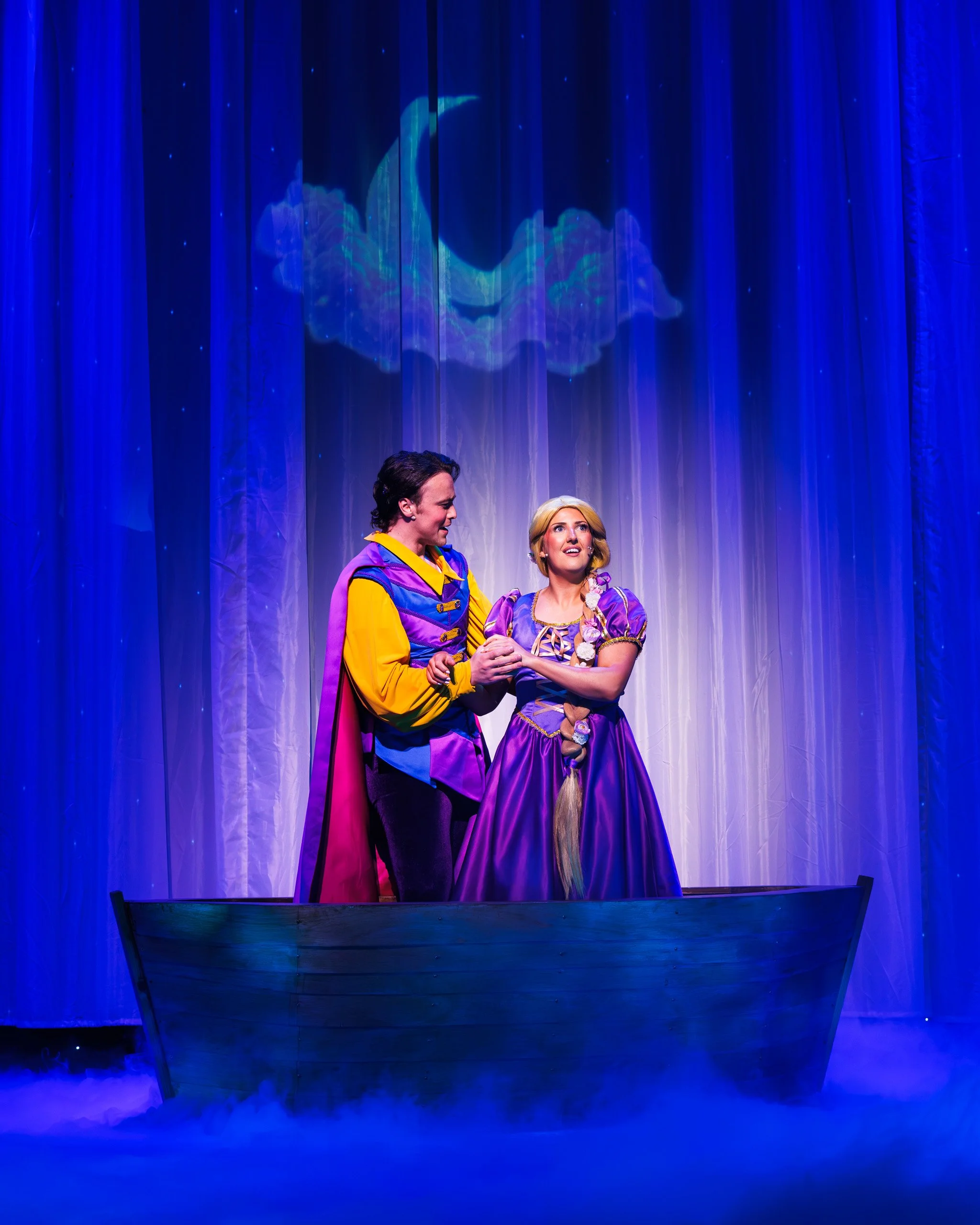 Two actors in colorful costumes on stage, one in a purple and yellow outfit with a cape, the other in a purple gown with a long braid, are holding hands and facing each other, with a blue curtain and a projection of a moon and clouds in the background.