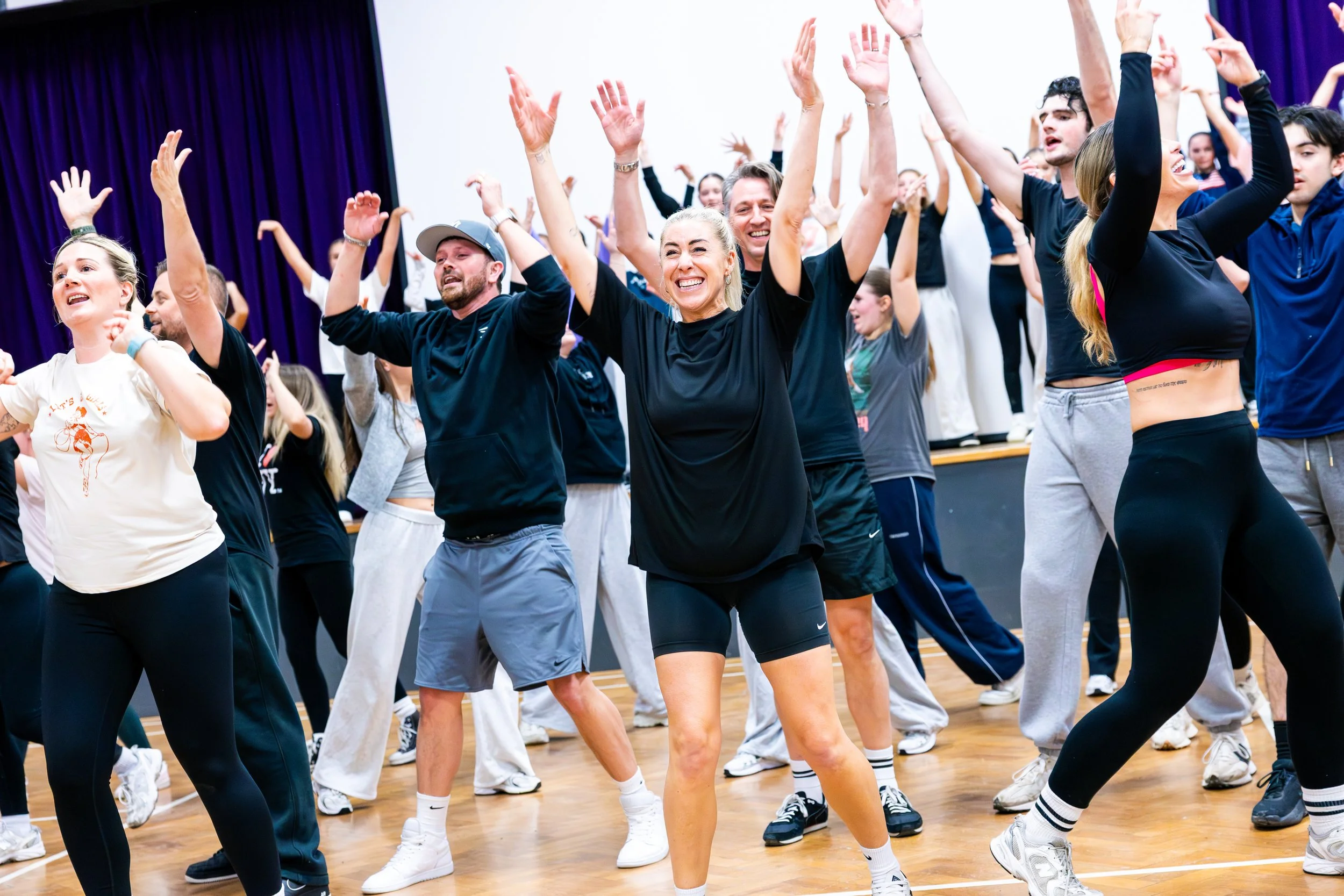 People dancing energetically in a gymnasium with a wooden floor, purple curtains, and white walls.