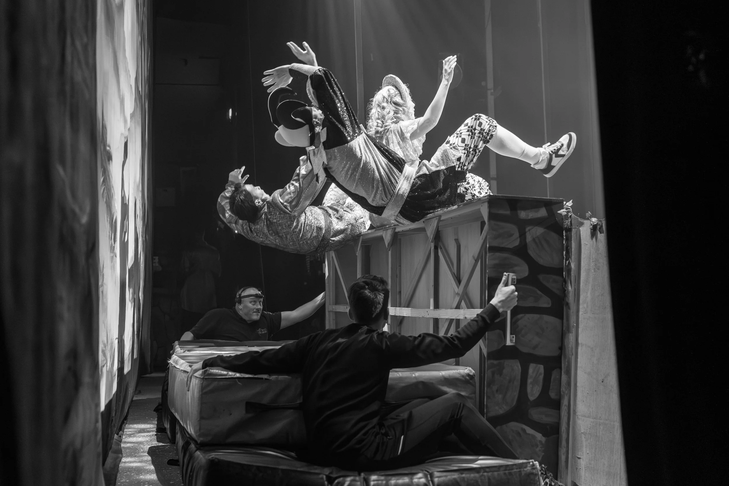 Black and white photo of a person lying on a padded surface, being lifted by a team of people, with one person below giving support. The scene appears to be part of a performance or stunt.