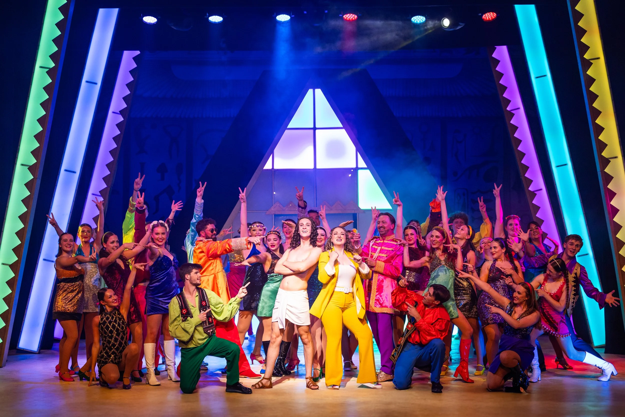 A large group of performers on stage in colorful costumes, smiling and posing with peace signs, under bright stage lights with neon border decorations behind them.