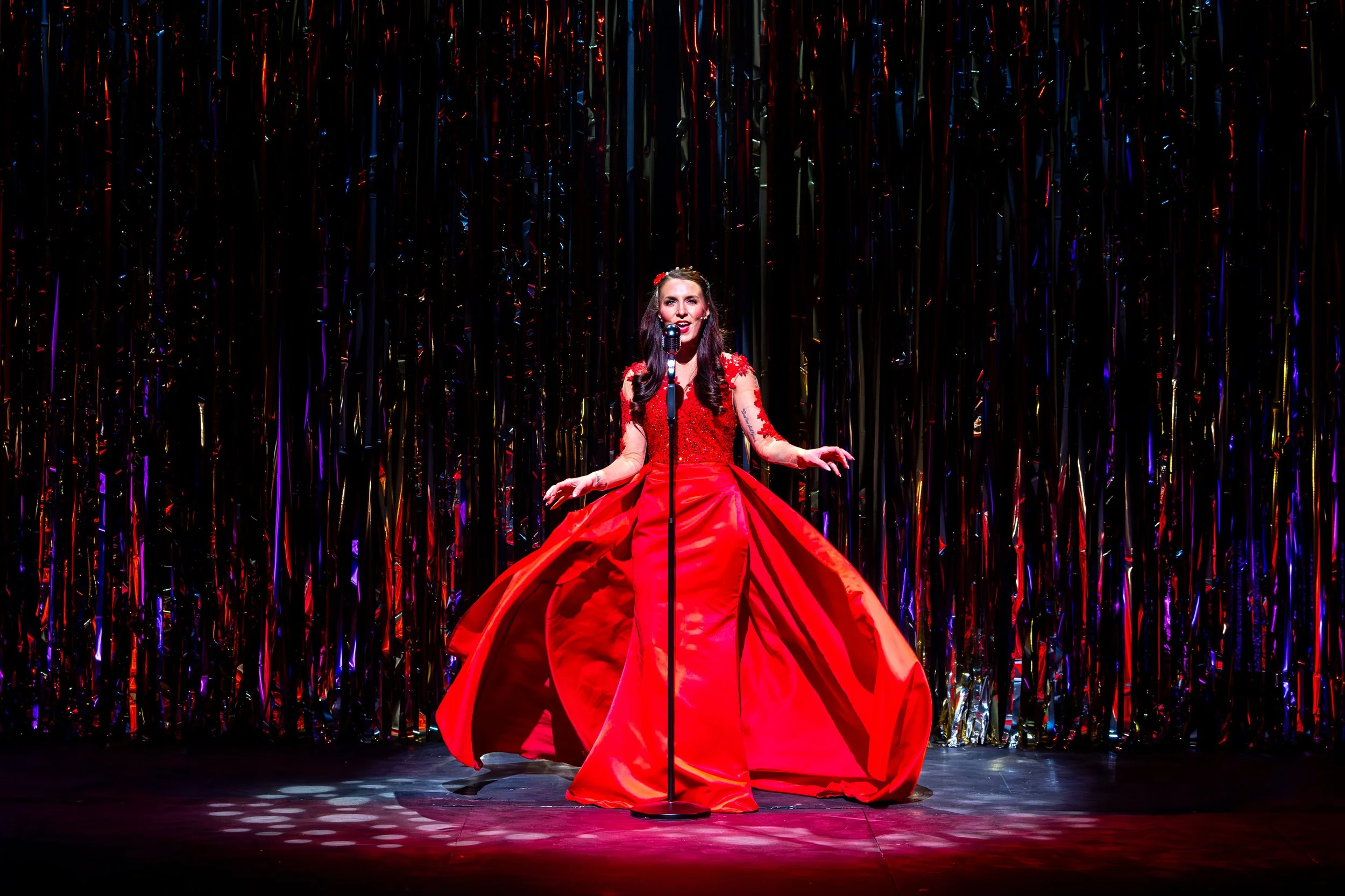 A woman in a red gown standing on stage behind a microphone with a dark background and colorful ribbons.