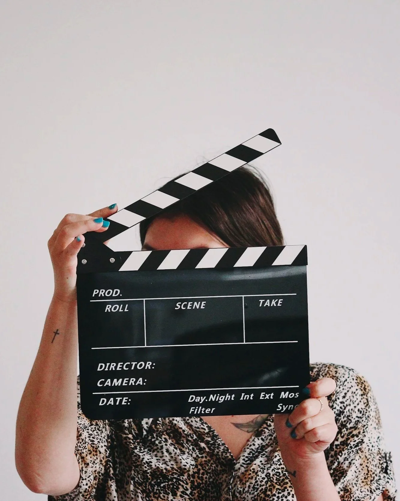 Person holding a clapperboard in front of their face with a white background. The person has short brown hair, wearing a leopard print top, and has teal-painted nails.