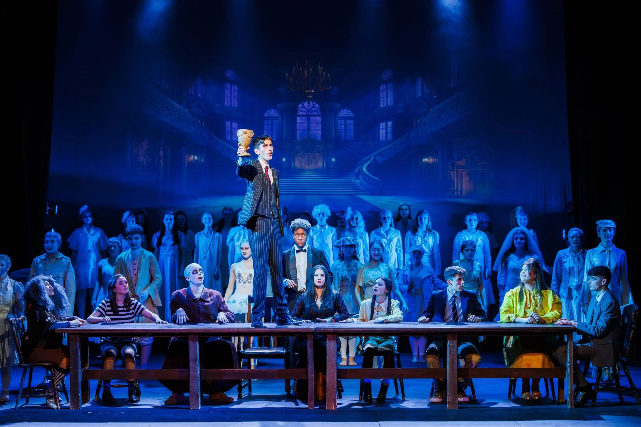 Theatre performance with a large cast of children and teenagers on stage, with a man in a striped suit holding a trophy, set against an elaborate mansion backdrop, lit with blue lighting.