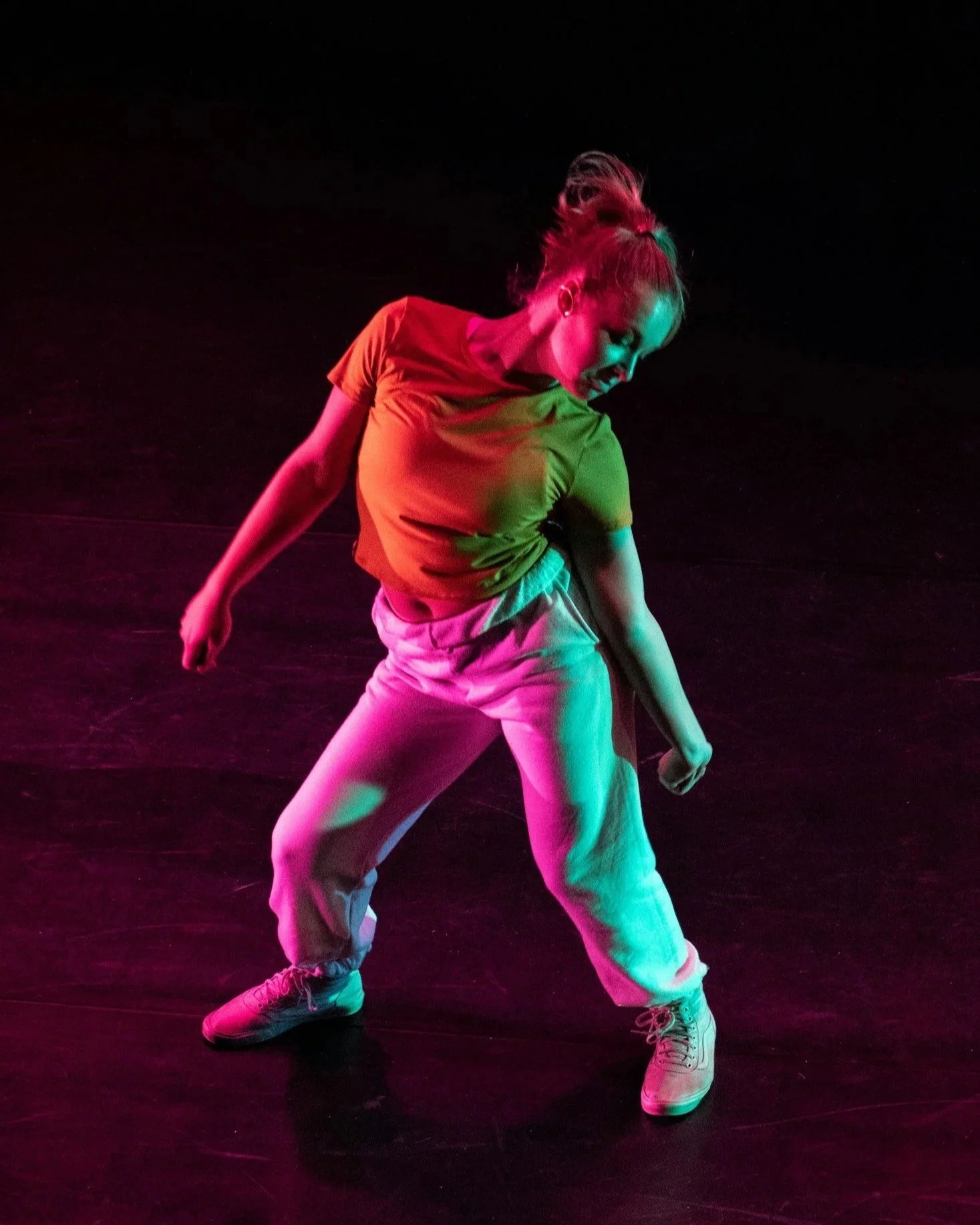 A woman with blonde hair styled in a high ponytail dancing on a dark stage, wearing a red t-shirt, light-colored pants, and sneakers, illuminated by colorful stage lights.
