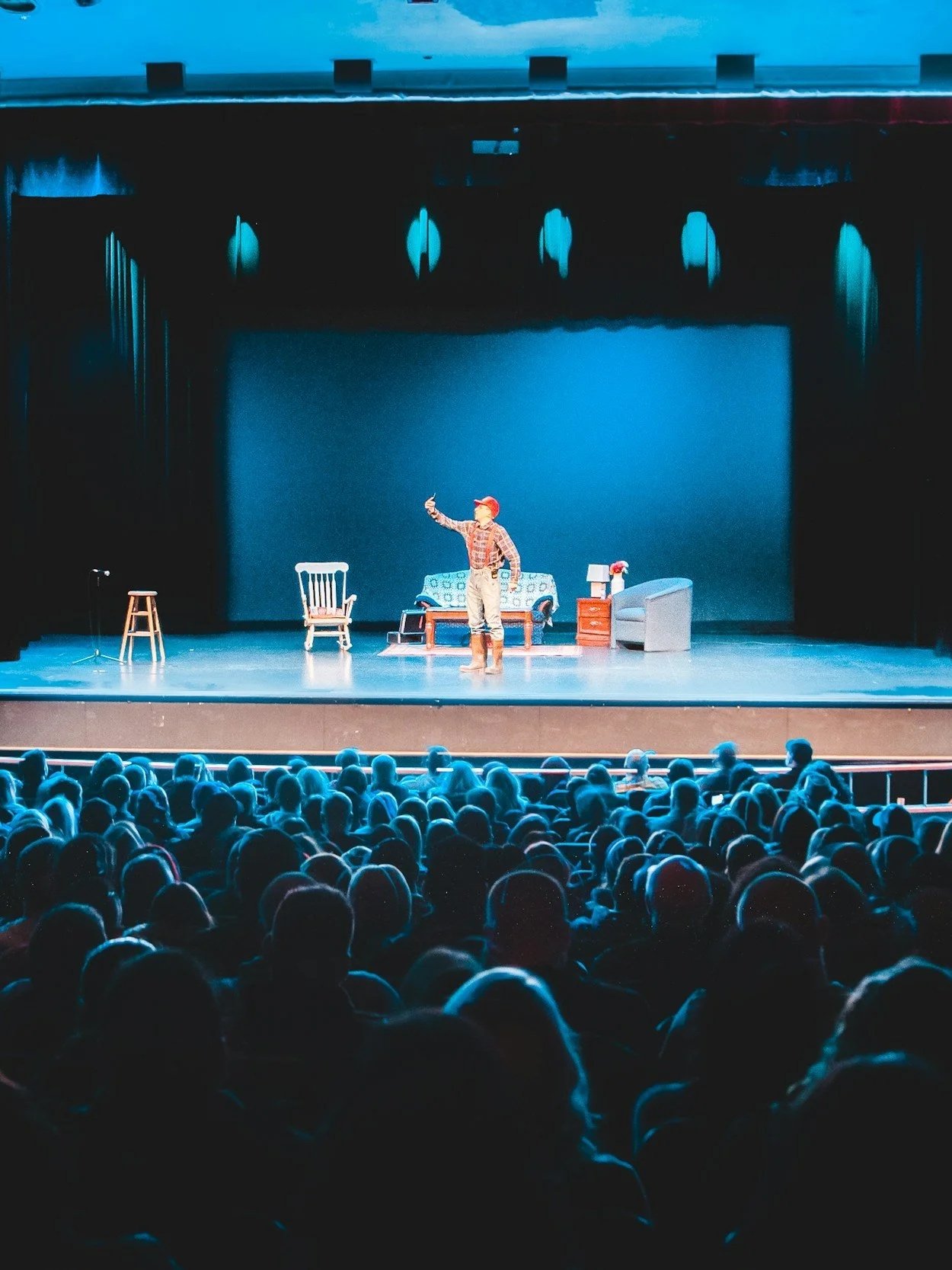 A stage with a performer dressed as a farmer or cowboy, taking a selfie with a smartphone, in front of a set of furniture including chairs, a sofa, and a side table with a lamp. An audience is seated watching the performance.