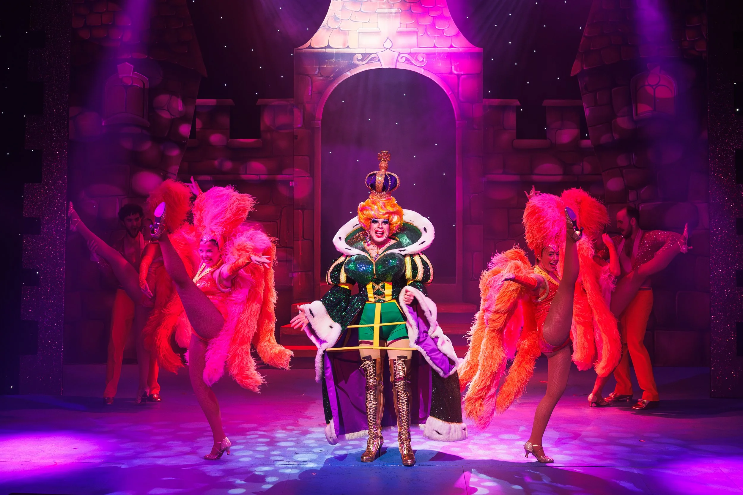 A theatrical stage scene with a performer dressed as a queen with a colorful, elaborate costume and crown, surrounded by dancers in bright orange and pink costumes with large feathered accessories, all under vibrant purple and pink lighting with a ca