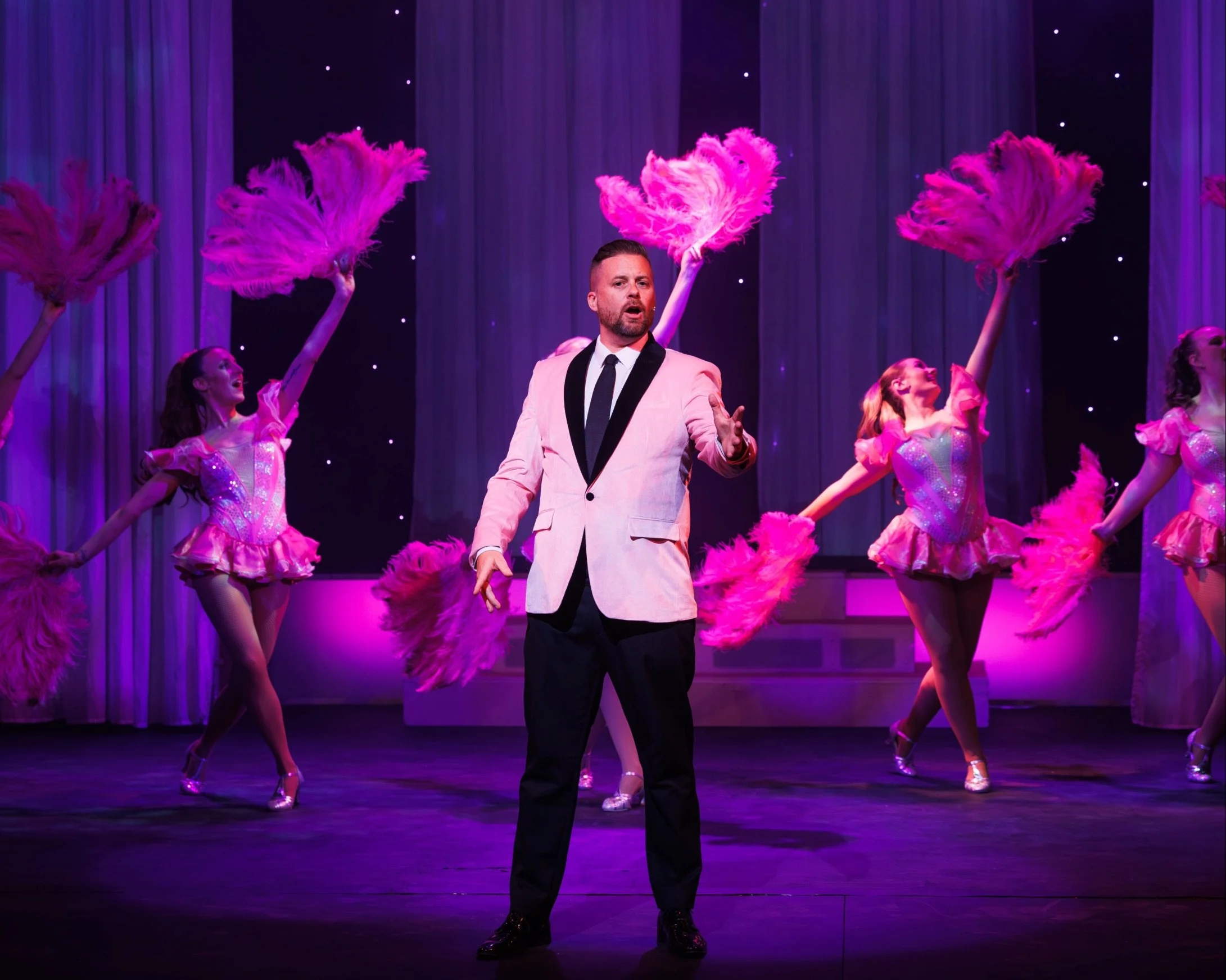 Man in a light pink suit jacket and black tie standing on stage with female dancers in pink costumes with feathers and pink fans, purple curtains background.