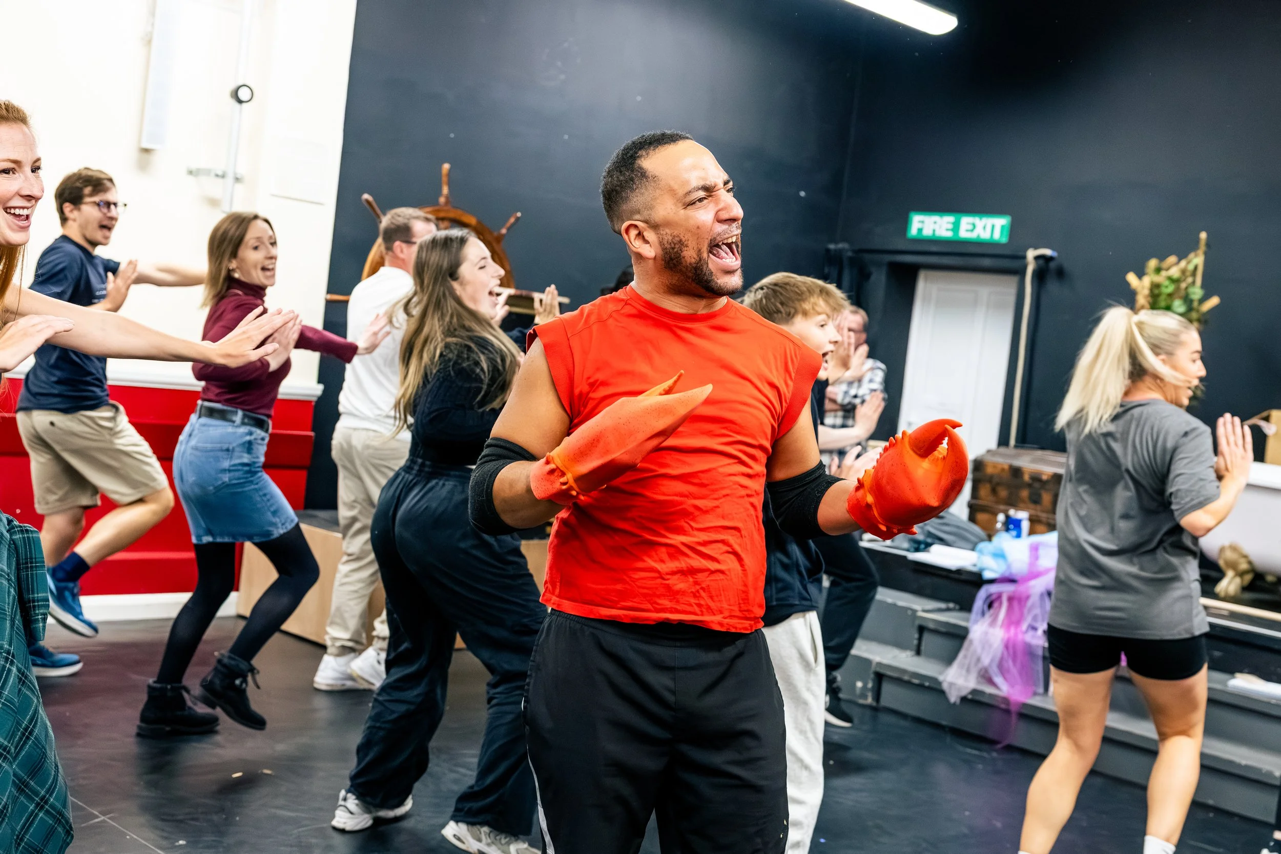 A group of people, including a man with boxing gloves, participating in a dance or exercise class indoors. They appear to be happy and energetic, with some smiling and reaching out. There is a dark wall with an exit sign and furniture in the backgrou