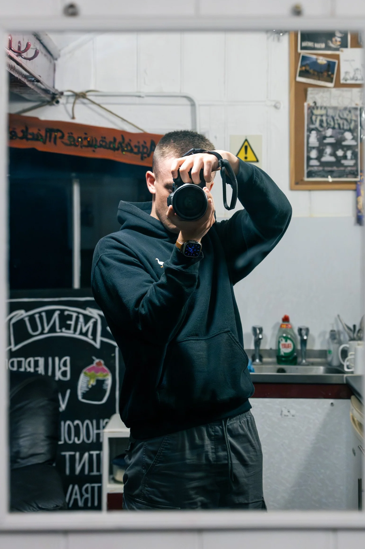 A young man taking a selfie in a mirror in a kitchen, holding a camera in one hand, wearing a black hoodie, with a short haircut and a smartwatch on his wrist.