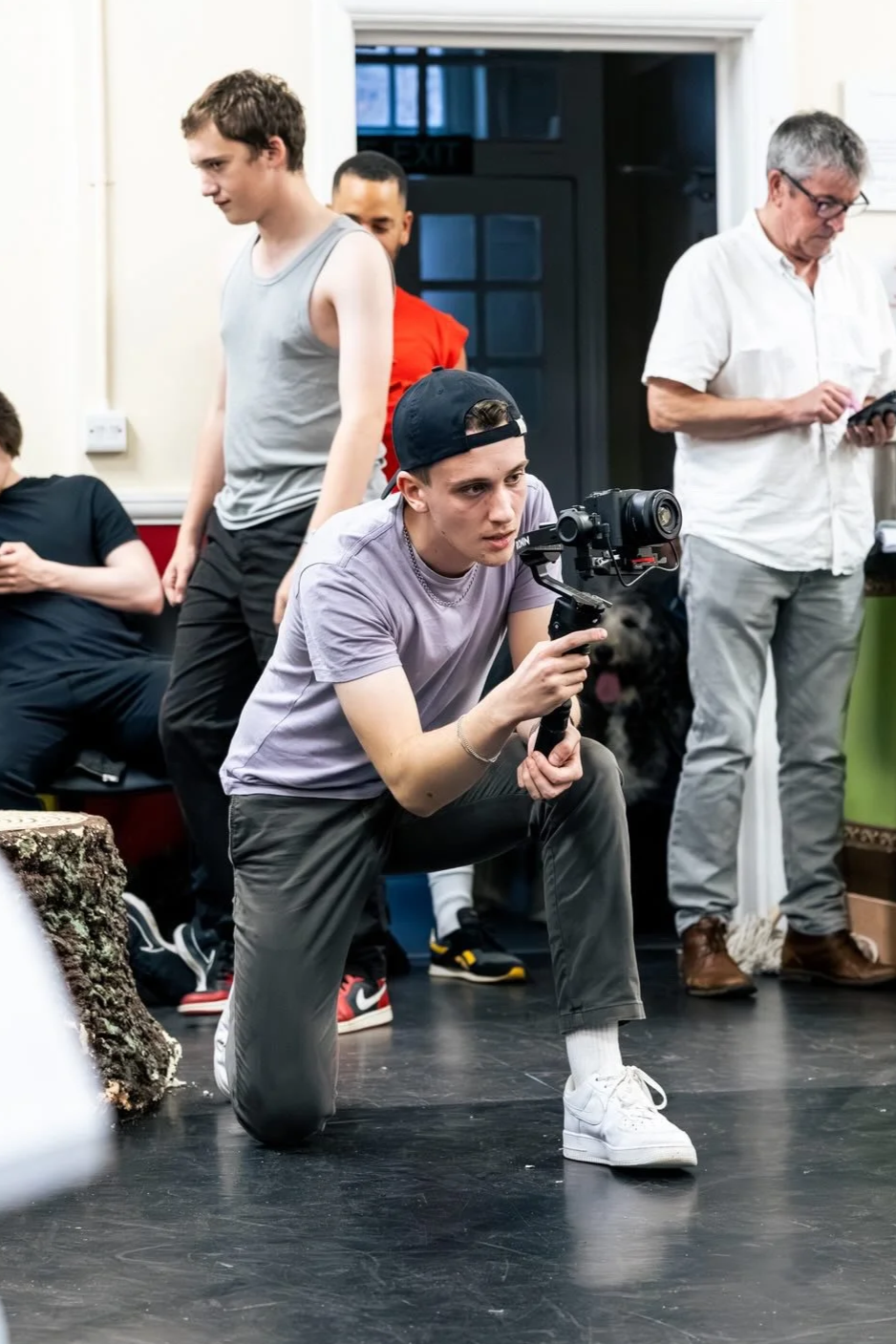 Young man wearing a black cap, gray t-shirt, and gray pants kneeling on one knee, holding a camera stabilizer with a camera, in a room with several people and a black dog in the background.
