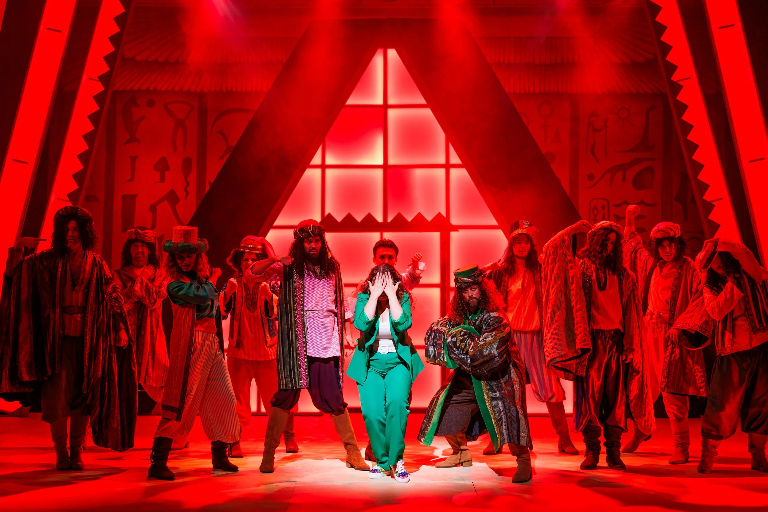Stage performance featuring a diverse group of actors dressed in colorful costumes with a red-lit background and large window, possibly from a musical or play.