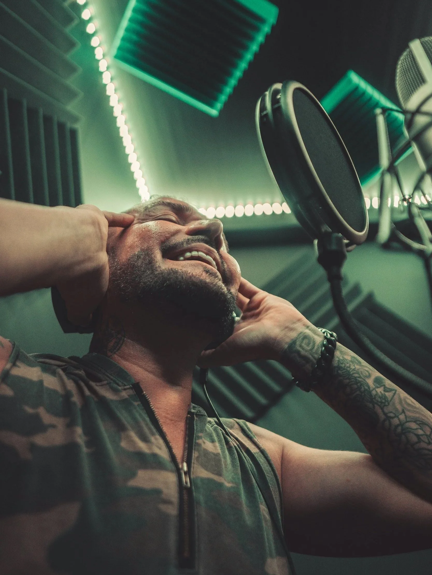 A man with tattoos on his arm, wearing a camouflage shirt, is smiling and holding his head while sitting in a recording studio. There is a microphone in front of him, and soundproofing foam panels and green LED strip lights are visible on the wall in the background.