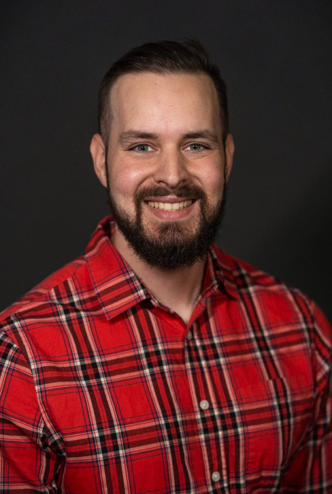 A smiling man with a beard and mustache wearing a red plaid shirt against a dark background.