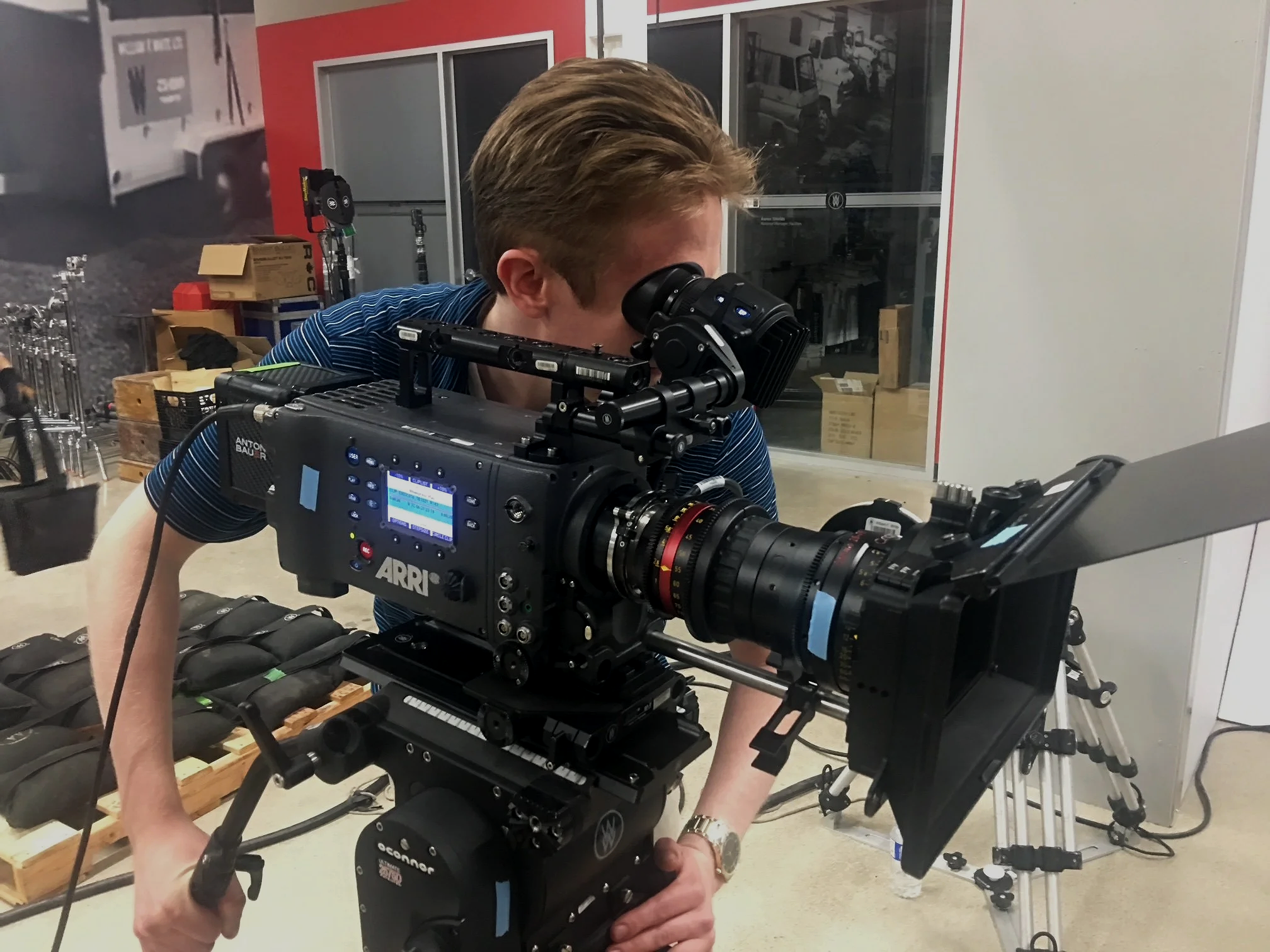 A man with light brown hair looking through a professional camera with a large lens, positioned indoors near a window with a warehouse or industrial area visible outside.