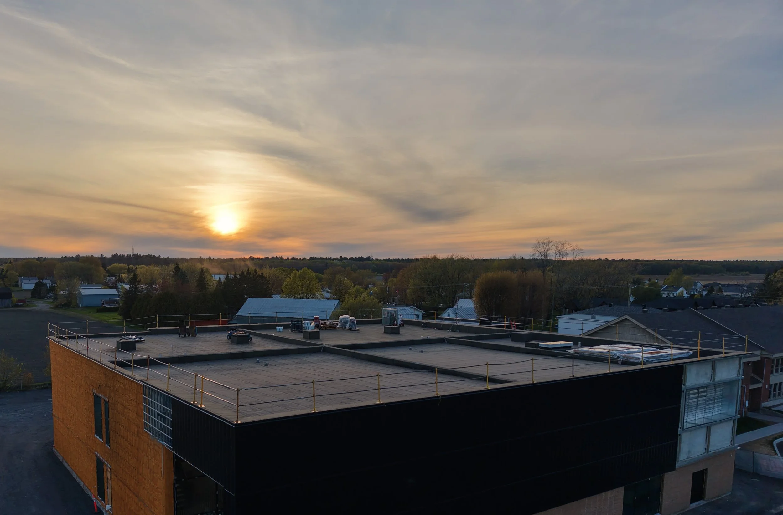 Un panorama d'un coucher de soleil derrière une étendue de bâtiments, avec des arbres au loin et un ciel avec des nuages légers.