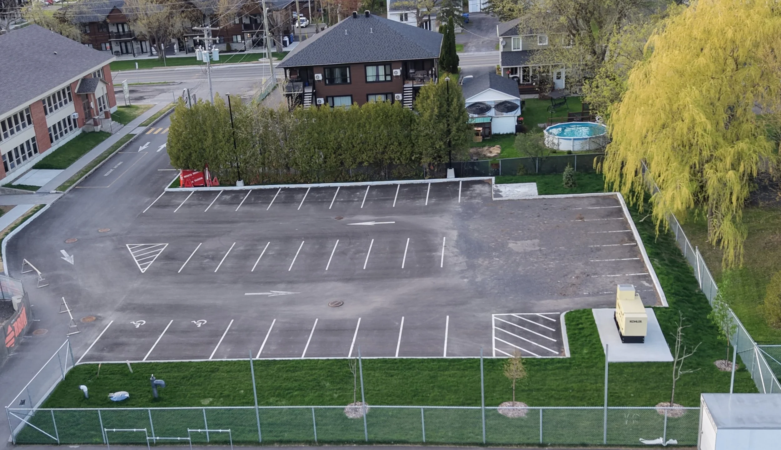 Photo d'un parking vide entouré d'une clôture, avec quelques bâtiments résidentiels et des arbres en toile de fond.