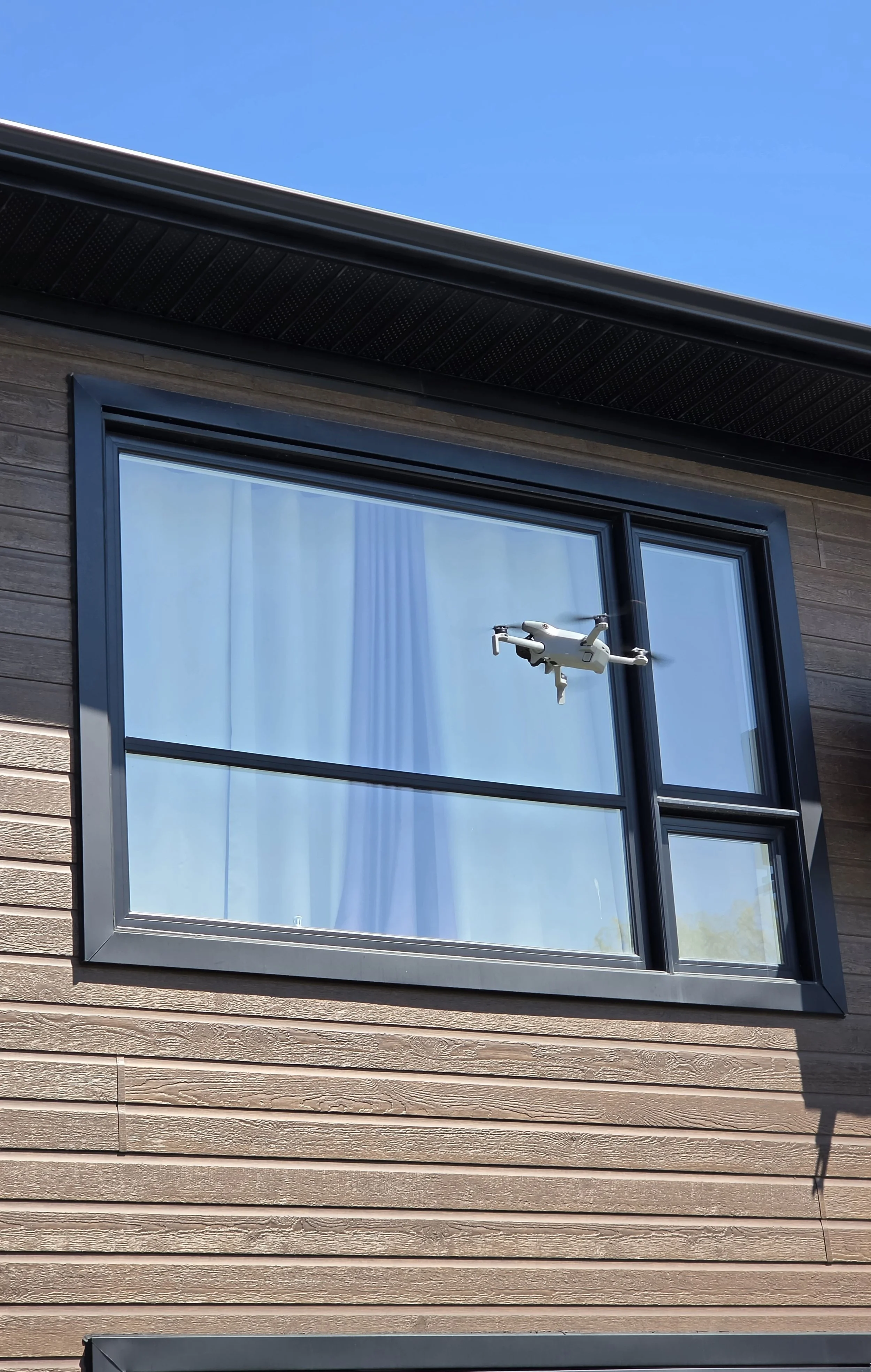 Une fenêtre de maison avec un drone à hover au centre, reflet de rideau blanc aperçu à travers la vitre, façade en bois. Ciel bleu en arrière-plan.