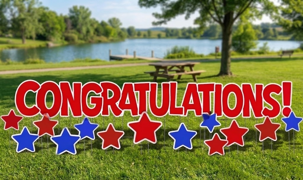 A colorful congratulatory sign with red, white, and blue stars in front of a lush park with a lake and picnic table.