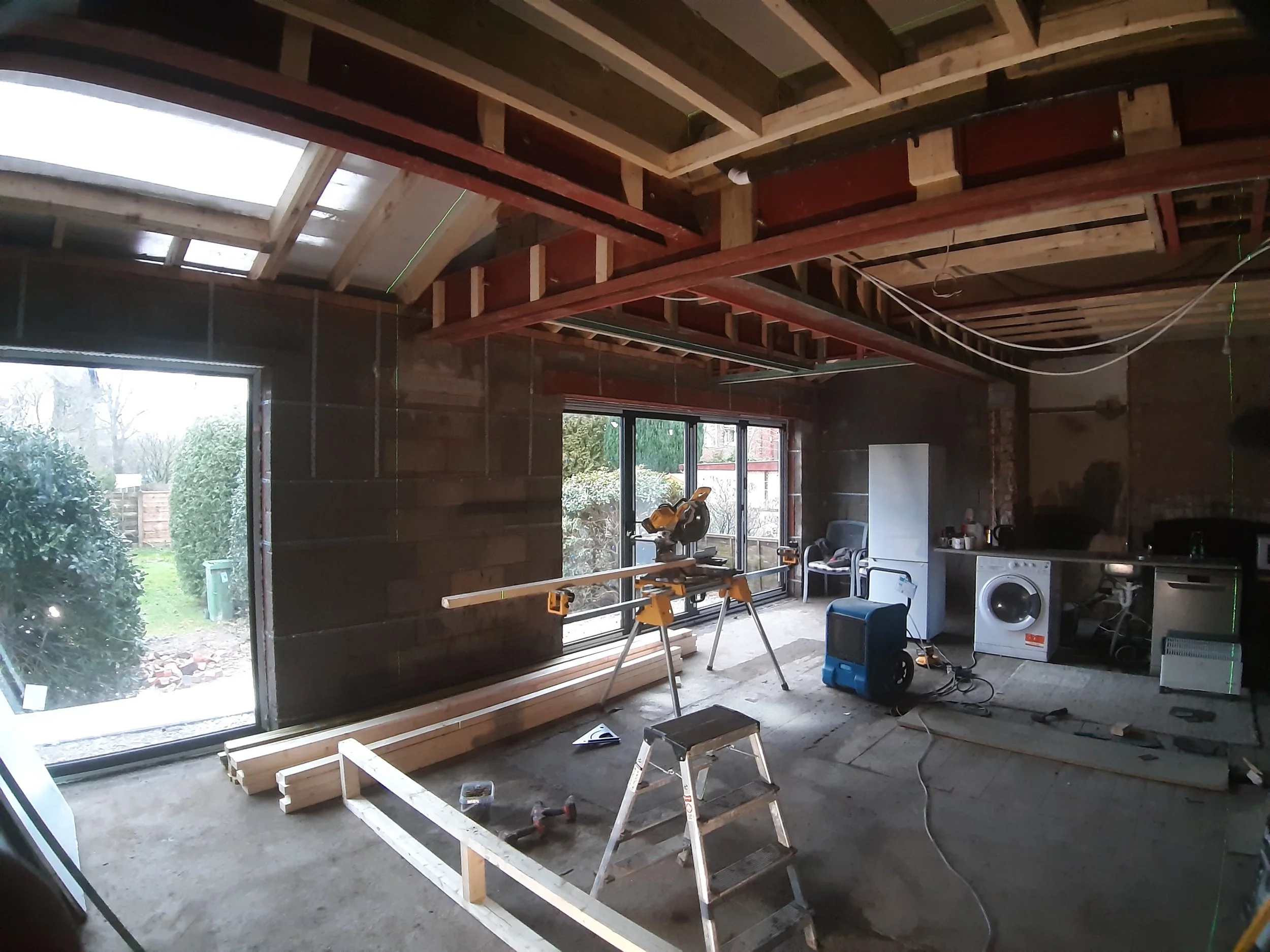 Interior of a house under construction with exposed ceiling beams, windows, and construction tools and materials inside, including wood planks, a saw, a step ladder, and electrical wires.