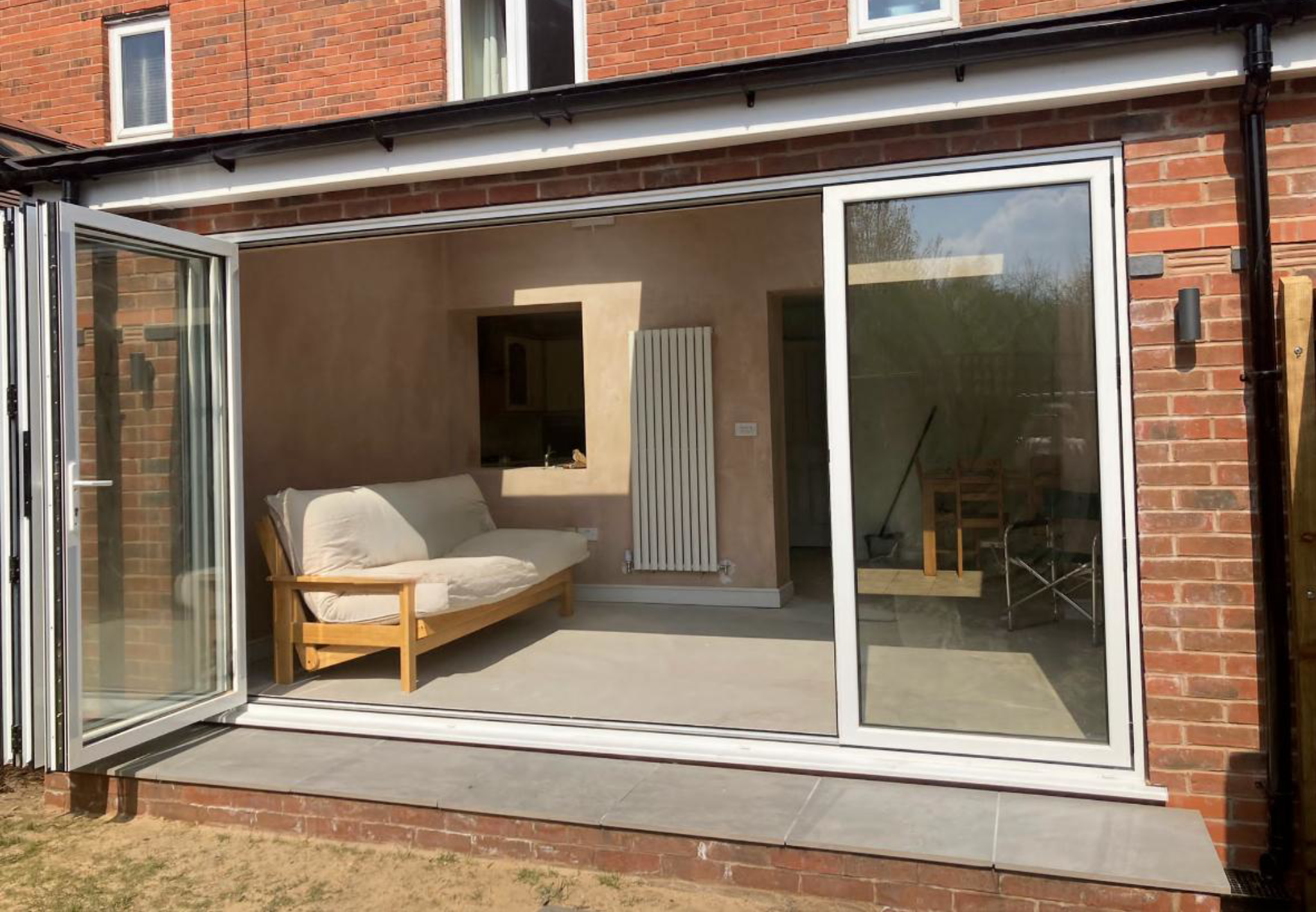 A brick house with an outdoor patio area featuring a sliding glass door. Inside, there is a beige hanging sofa with armrests and an open kitchen area with a window, radiator, and wooden chair.