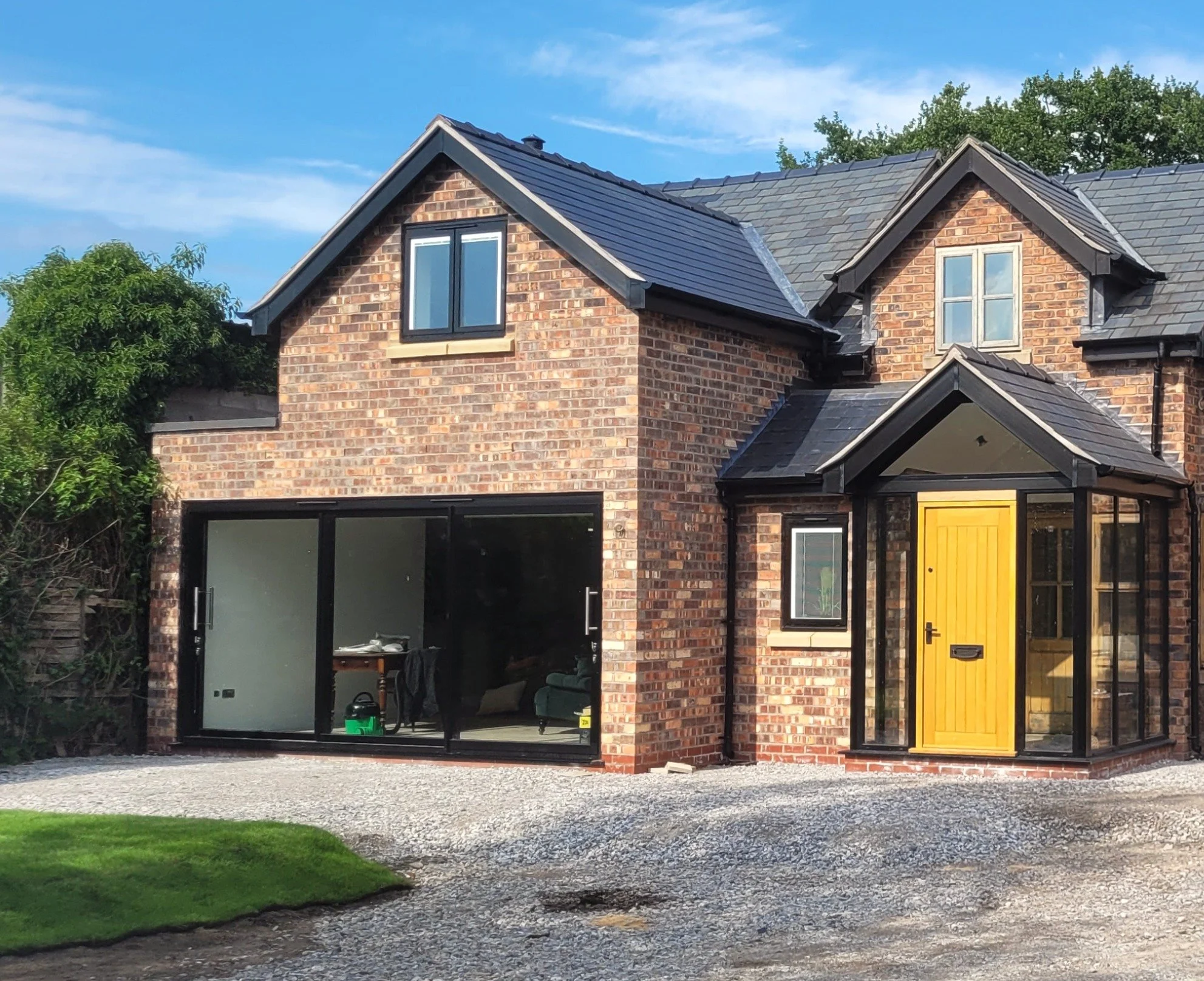 A brick house with a black and gray tiled roof and two gabled windows, one with black framing and the other with white framing, and a bright yellow front door, with a gravel driveway and a patch of green lawn in front.