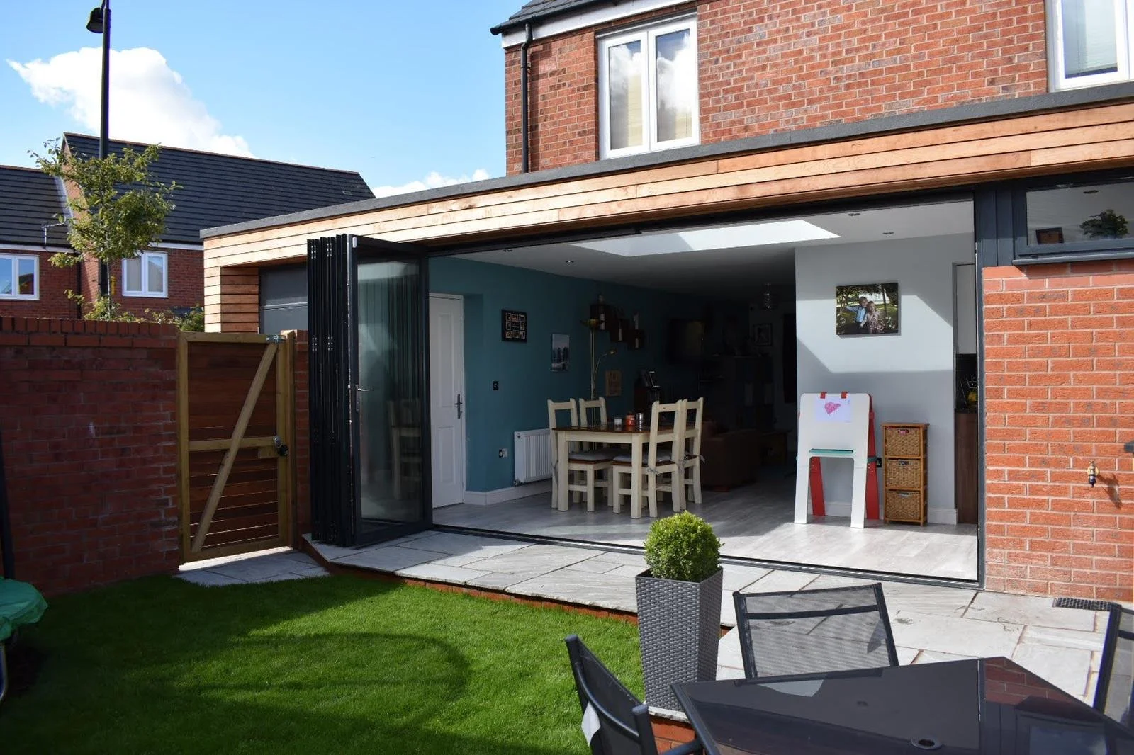 Open glass doors in a brick house reveal a dining area with a wooden table and chairs, inside a modern living space with blue walls, a white radiator, and various decorations. The outdoor area features a small lawn, a patio with chairs, and a potted plant.