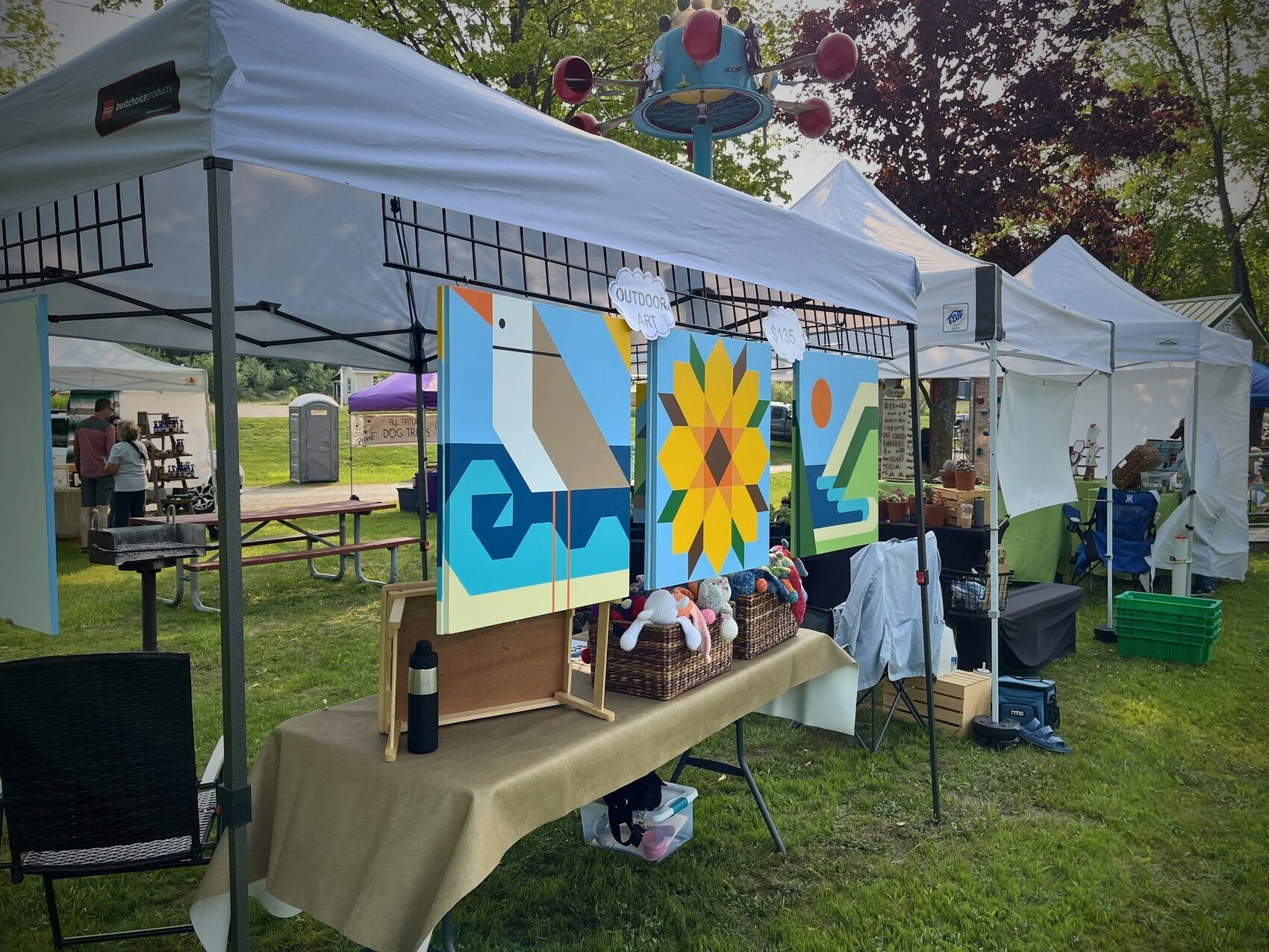 Photograph of an outdoor booth at a fair displaying three hanging barn quilts.