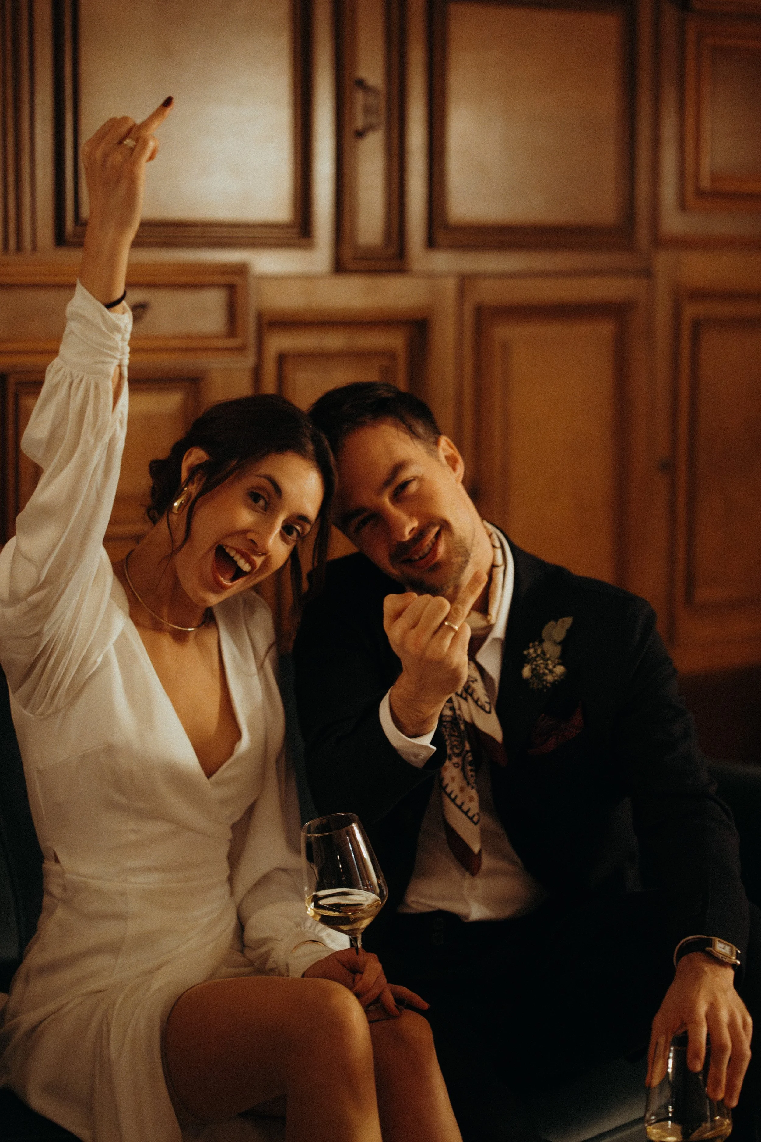 Zwei feiernde Menschen, ein Mann und eine Frau, Hochzeitspaar,  sitzen in einem Raum mit Holzwänden und trinken Wein. Die Frau trägt ein Hochzeitskleid und der Mann trägt einen schwarzen Hochzeitsanzug in Berlin.