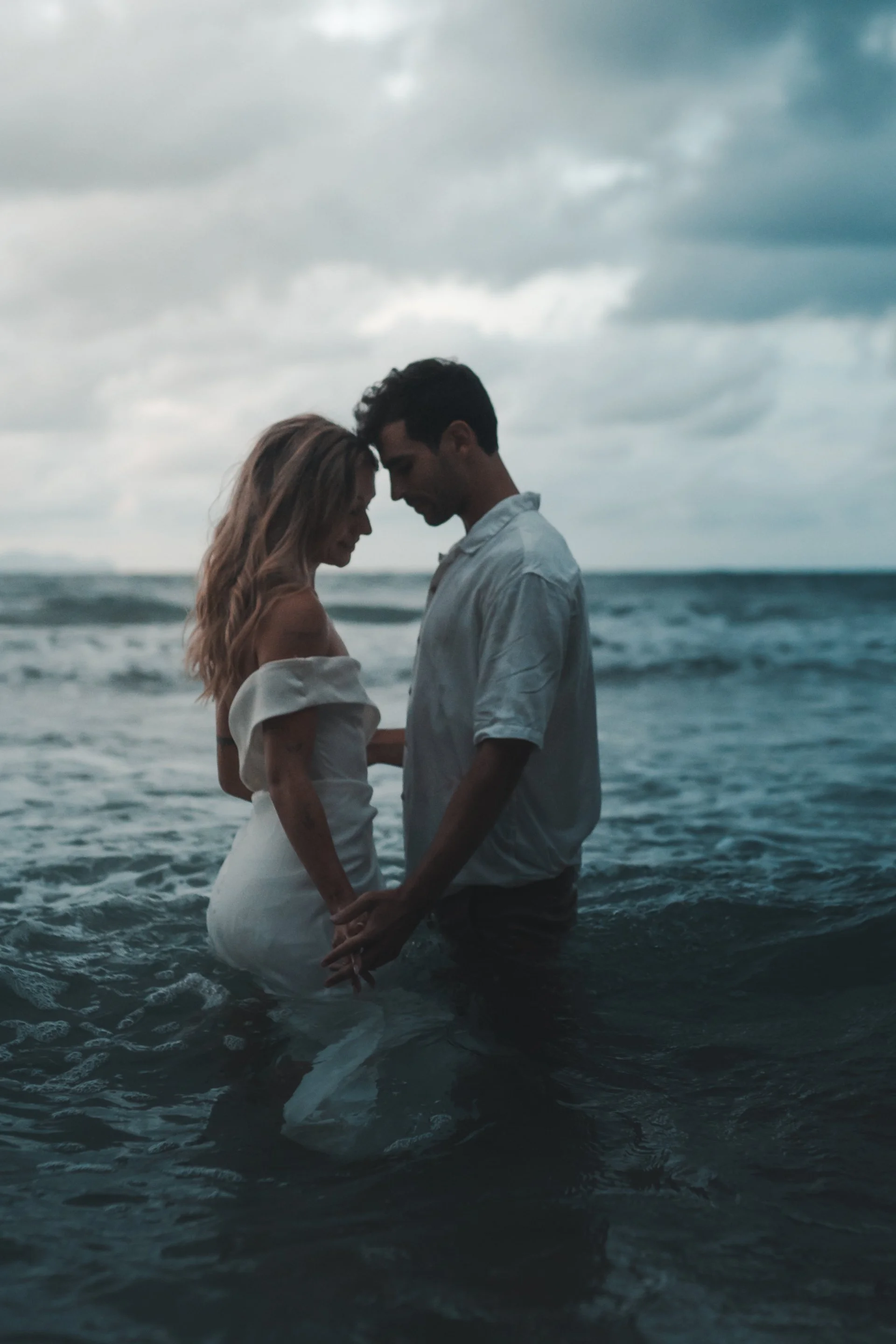 Ein Hochzeitspaar steht im Meer, berühren sich mit den Stirnen, während die Wellen um sie herum schlagen. Der Himmel ist bewölkt und die Stimmung ist romantisch und intim.