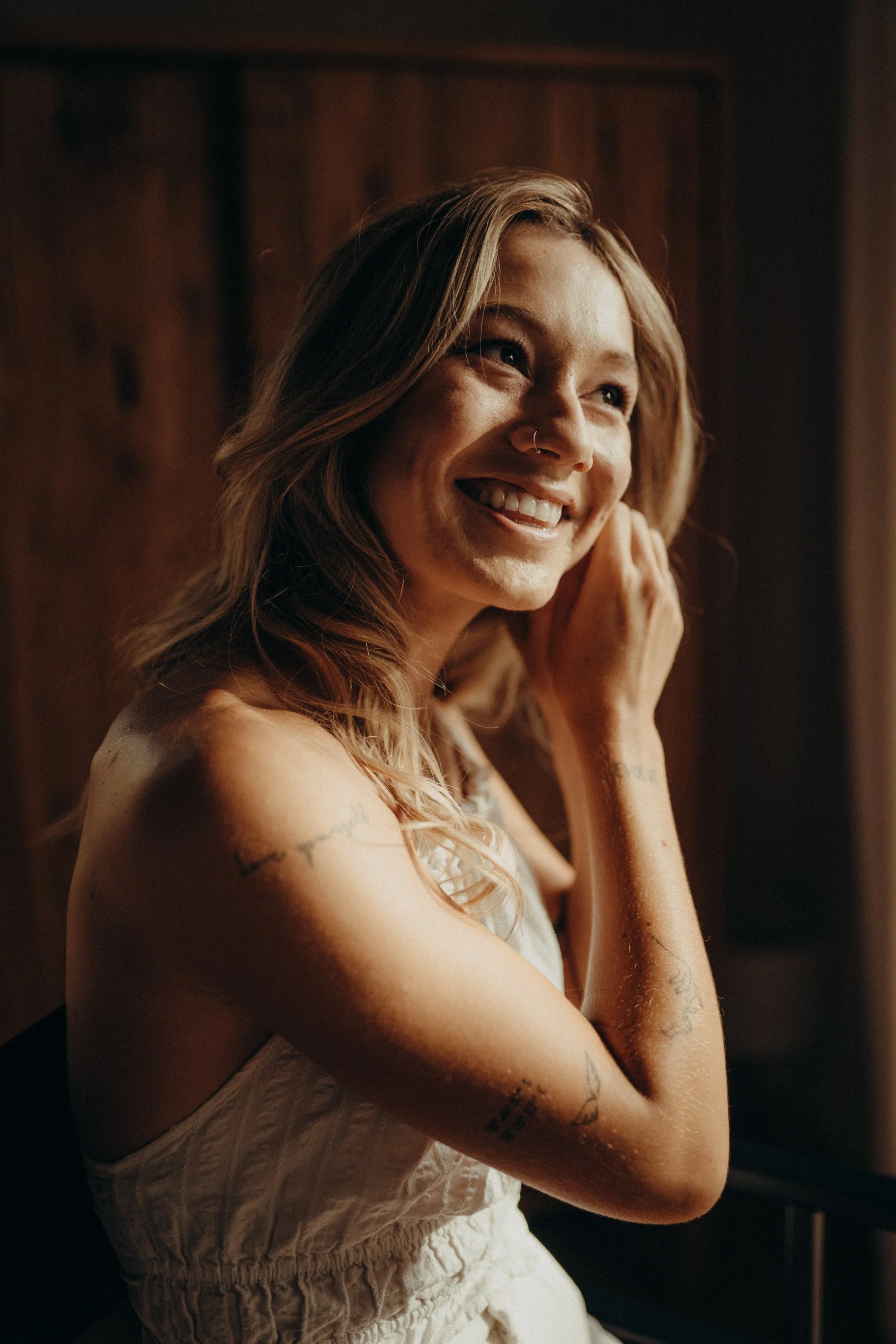 Lächelnde junge Braut mit Tattoos am Tag ihrer Hochzeit, während ihres Getting Ready, trägt ein weißes Kleid, hat blonde Haare und trägt einen Nasenring.