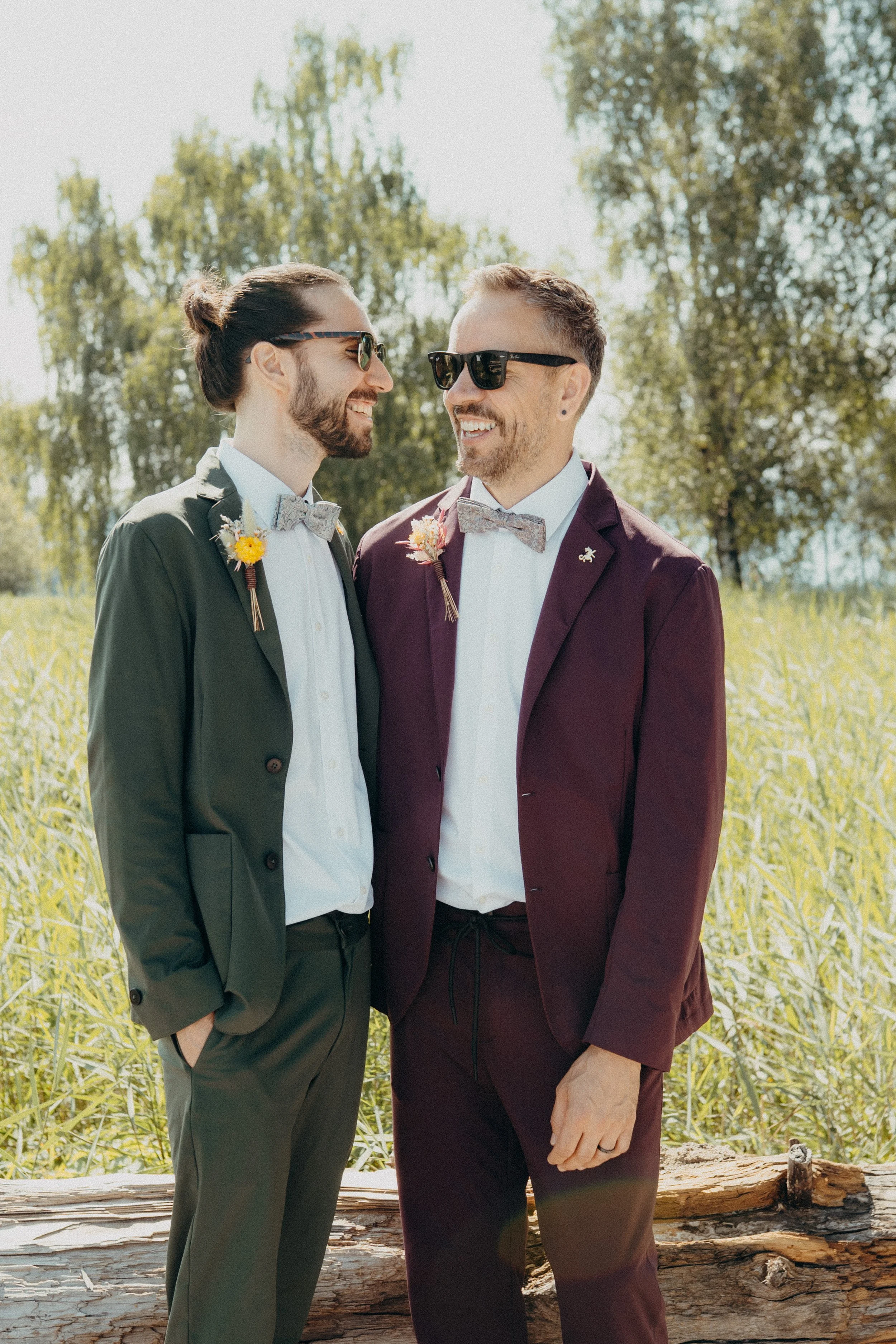 Hochzeitspaar - Zwei Männer kurz vor ihrer Hochzeit in Anzügen und Sonnenbrillen stehen in einer grünen Wiese im Freien, lachen und schauen sich an.
