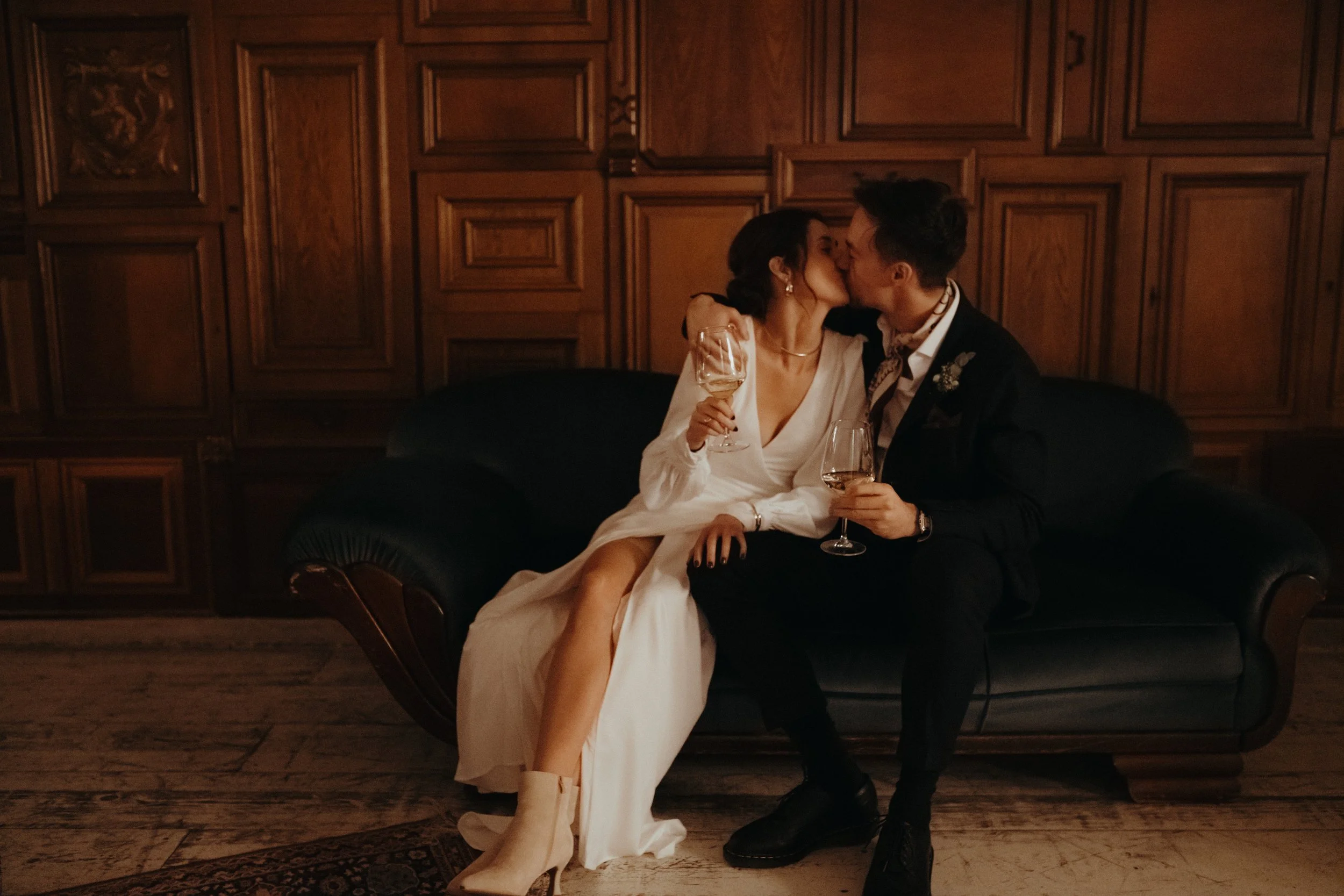 Ein Hochzeitspaar küsst sich auf einer Couch in einem Raum mit dunklen Holzwänden. Sie hält ein Weinglas, er trägt einen Hochzeitsanzug mit einem Halstuch.