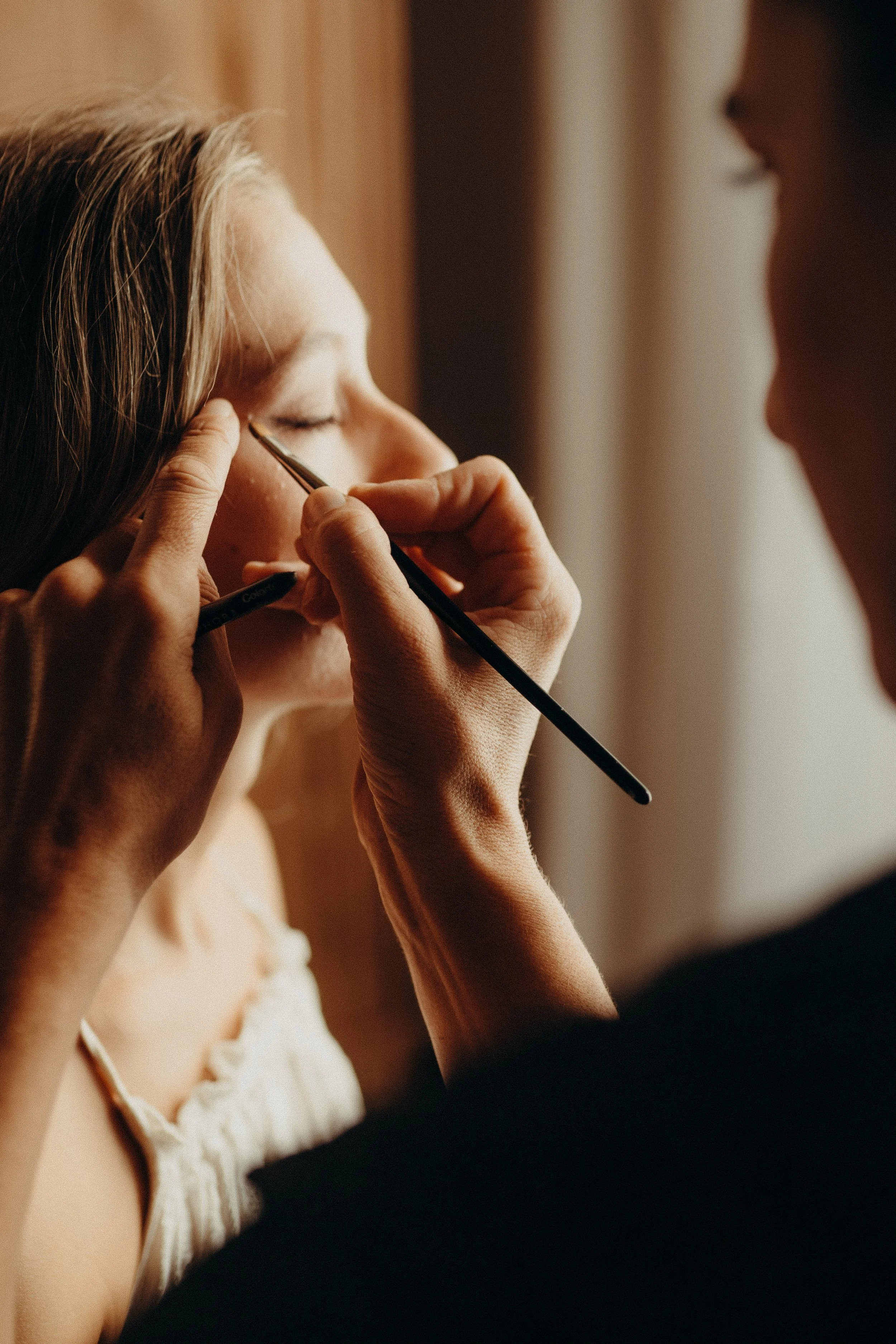 Jemand macht eine Make-up-Anwendung an einer Frau, die geschlossen die Augen hat.