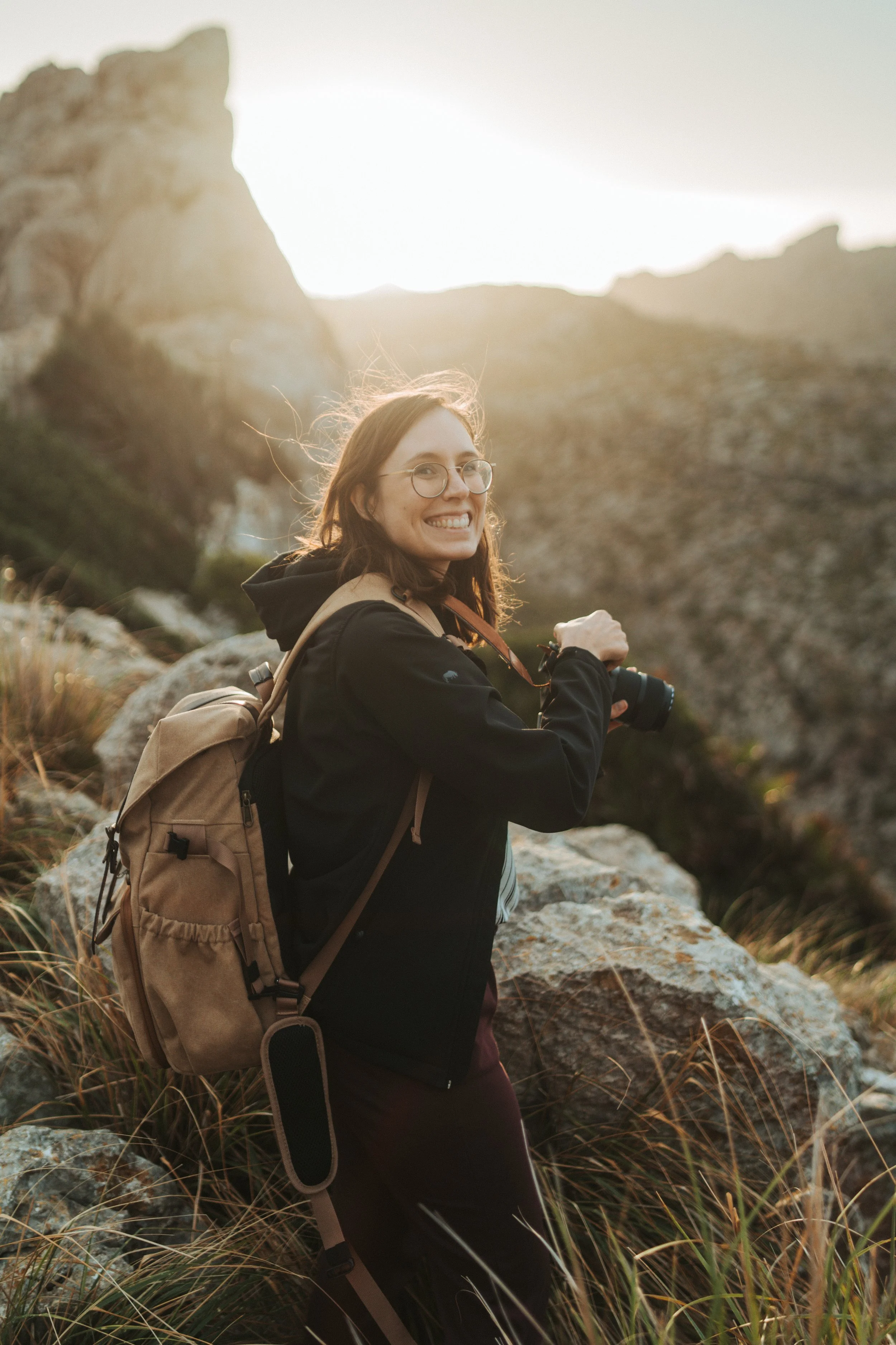 Eine lächelnde Frau mit Brille, Rucksack und Kamera beim Wandern in einer bergigen Landschaft bei Sonnenuntergang.