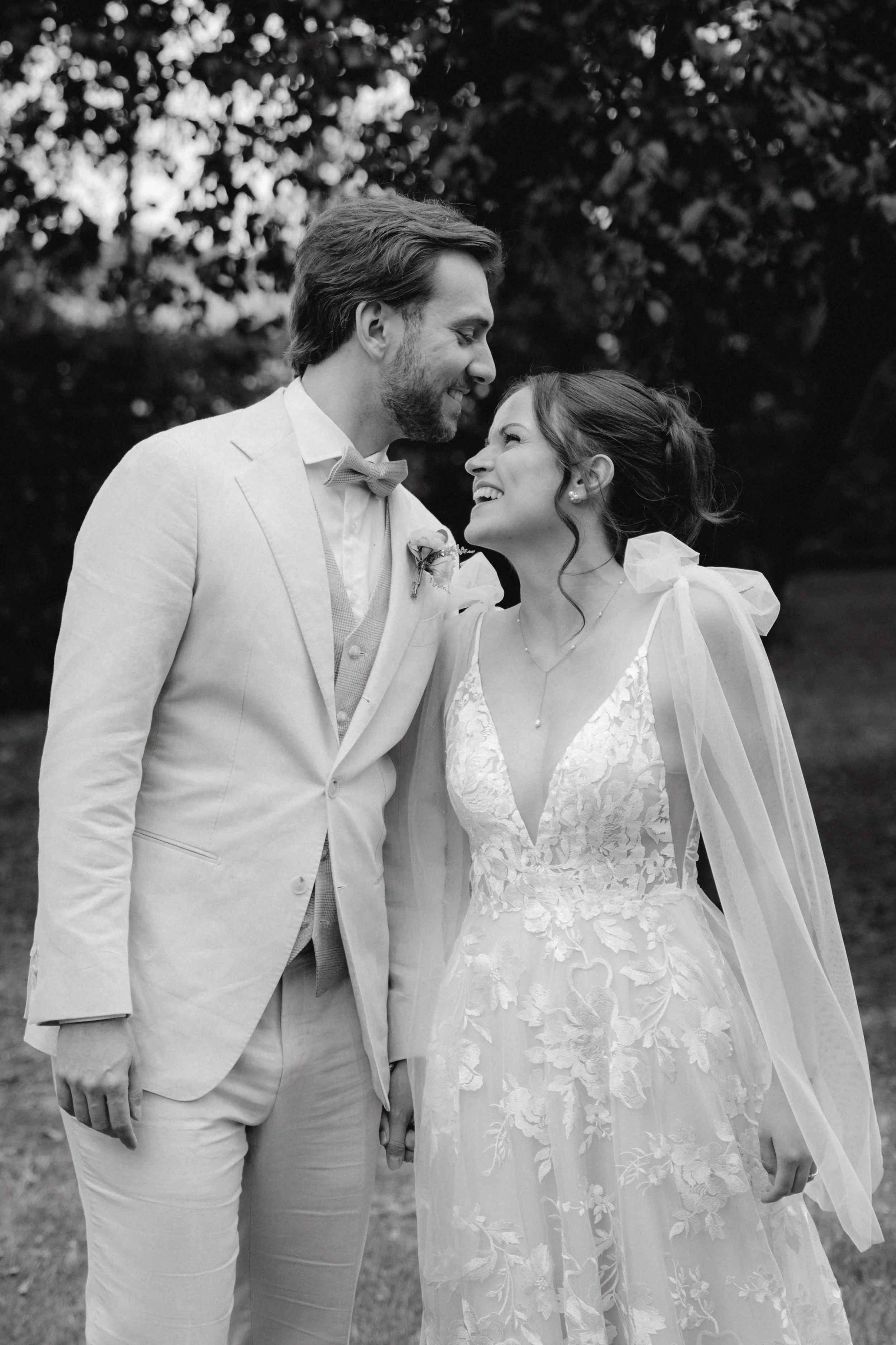 Ein Hochzeitspaar am Tag ihrer Hochzeit hält Händchen und lächelt sich liebevoll an, im Hintergrund Bäume, schwarz-weiß Fotografie.