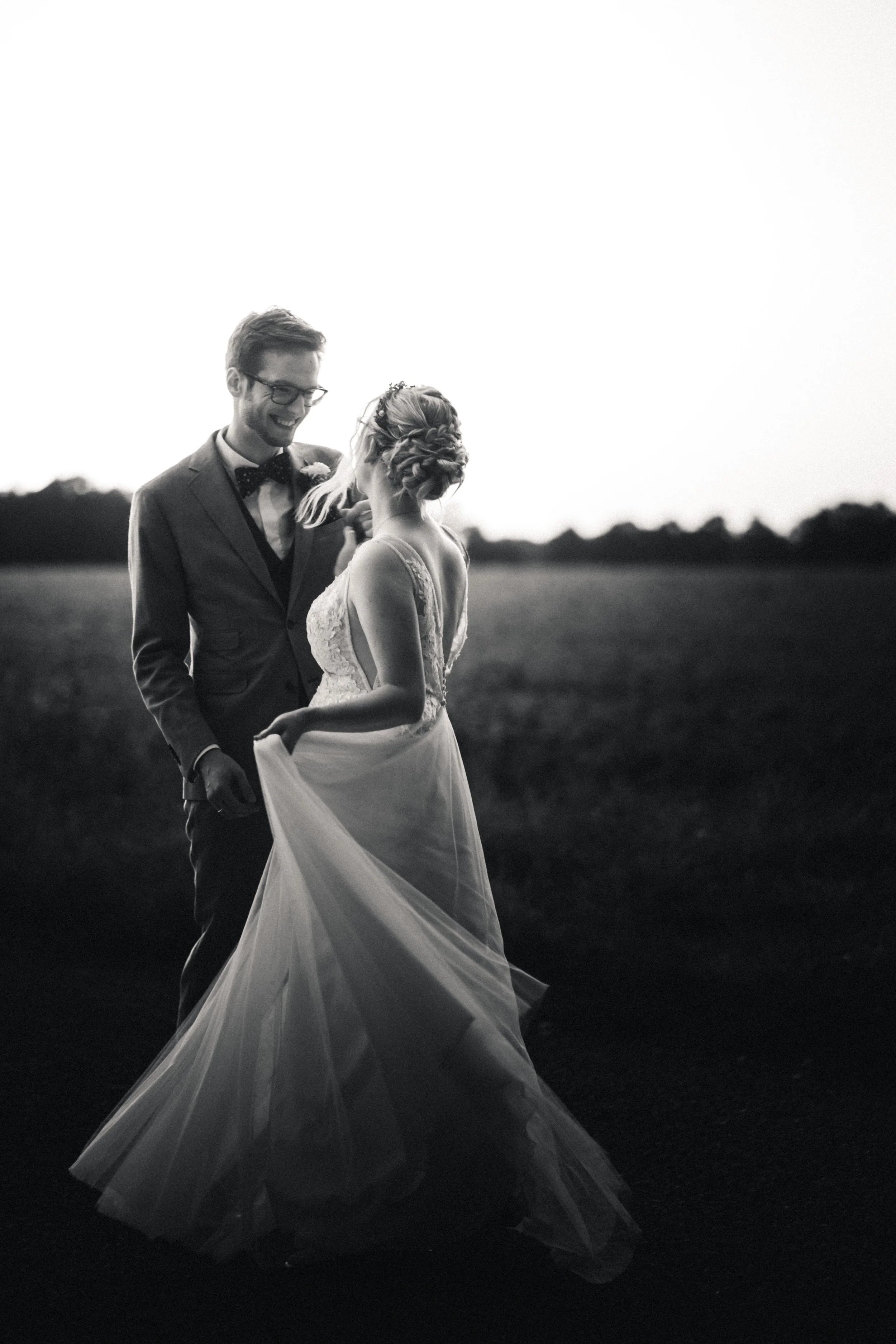 Ein Hochzeitspaar tanzt im Freien bei Sonnenuntergang, der Mann trägt einen Anzug mit Fliege, die Frau trägt ein langes Brautkleid. Die Szene ist in Schwarz-Weiß und romantisch.