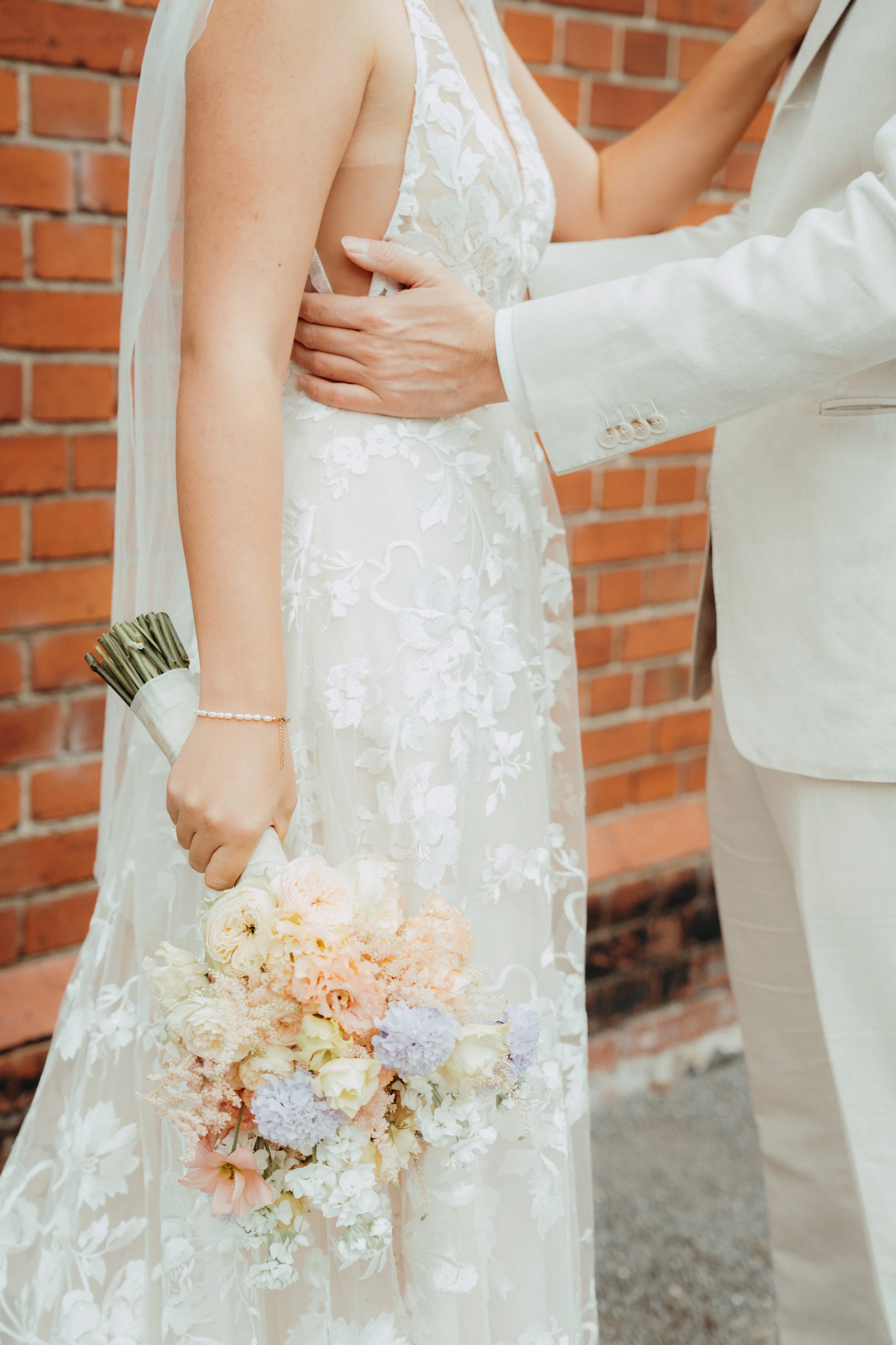 Ein Brautpaar bei einer Hochzeit, die Braut trägt ein weißes hochzeitskleid mit Stickereien, hält einen Blumenstrauß. Der Bräutigam trägt einen cremefarbenen Anzug. Sie stehen vor einer roten Backsteinmauer.