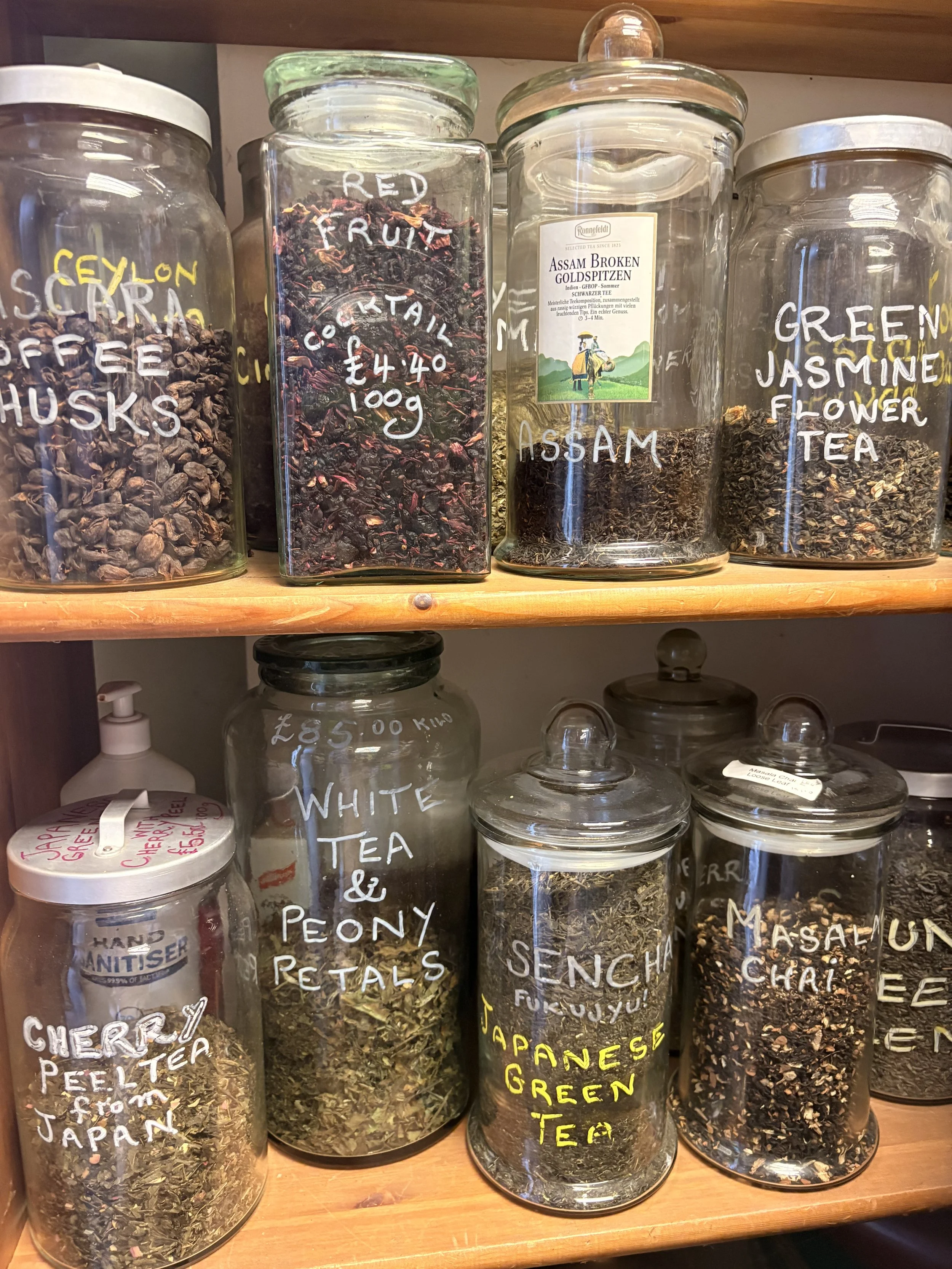 Clear glass jars filled with various loose teas, labeled with handwritten descriptions such as 'Seylon', 'Red Fruit', 'Assam Broken Goldspitzen', 'Green Jasmine Flower', and others, arranged on a wooden shelf.