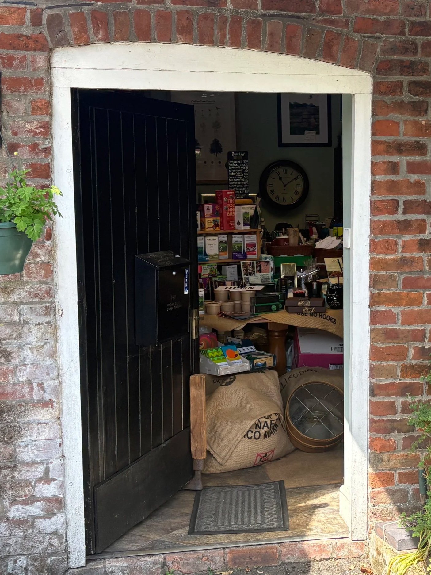 View of a small storefront or cafe entrance with a black door partially open, brick walls, and a white-painted arch. Inside, shelves display various packaged products, a clock on the wall, and bags and baskets on the floor near the doorway.