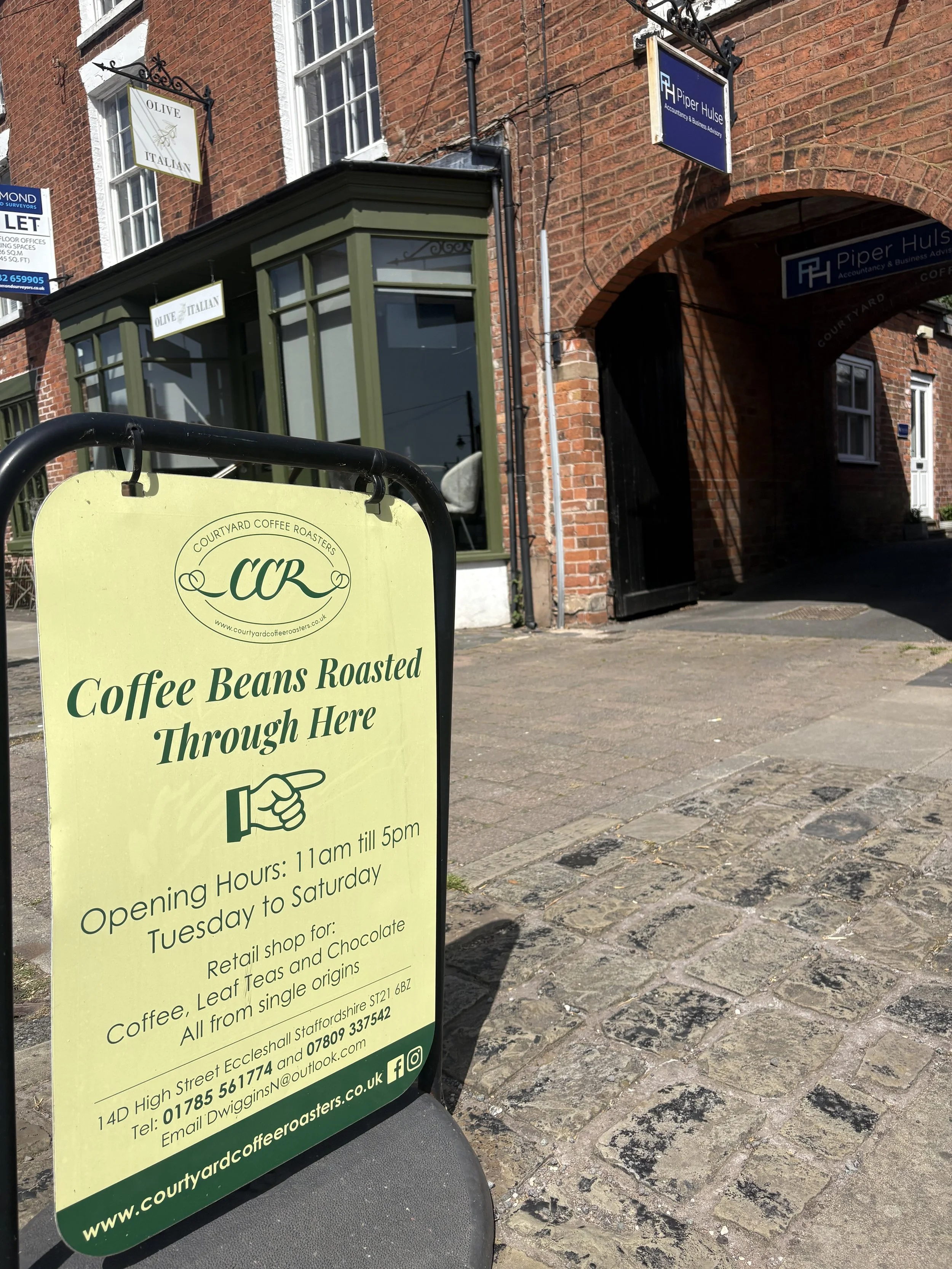 A yellow sandwich board sign outside a coffee shop advertises coffee beans roasted on site, with shop hours and contact information. The background shows a brick building with windows and signs for other businesses.