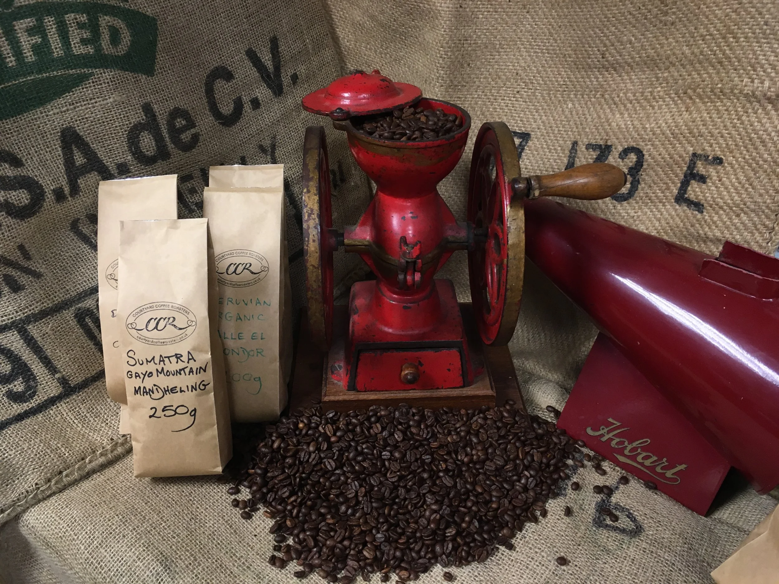 Vintage red coffee grinder with coffee beans spilled in front of it on a burlap background, with paper bags of coffee beans nearby.