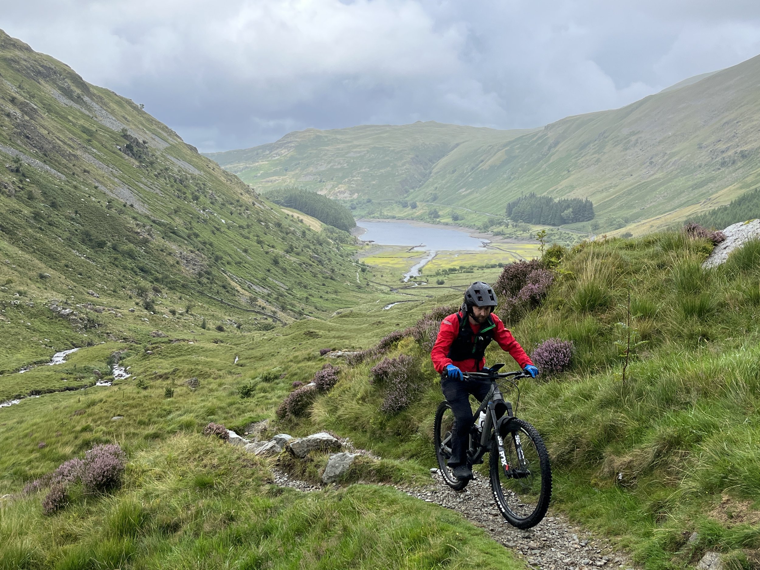 A person wearing a red jacket, helmet, and gloves riding an electric mountain bike on a narrow dirt trail in a lush green valley surrounded by hills and mountains, with a lake visible in the distance under a cloudy sky.