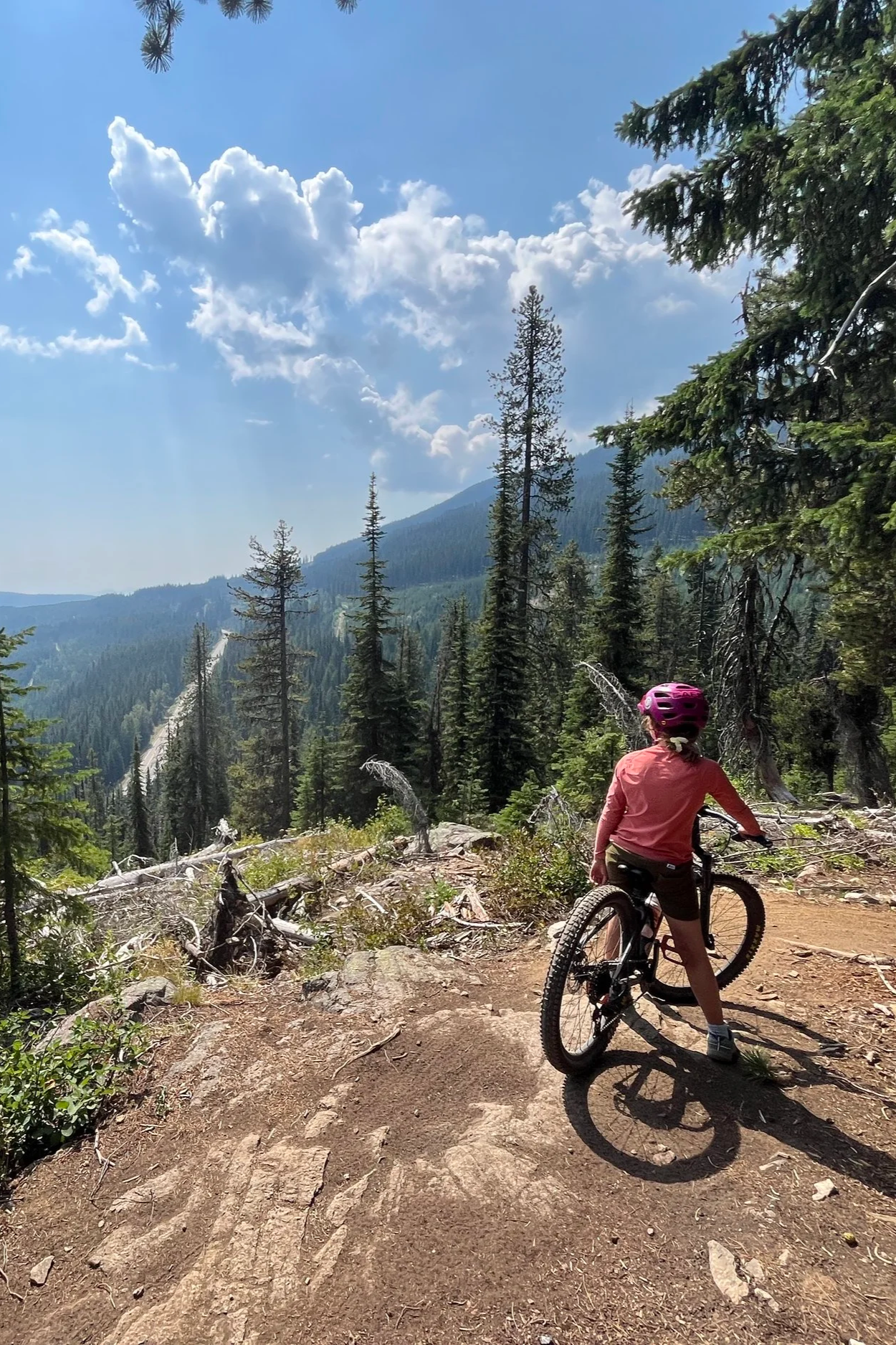 A person wearing a pink helmet and pink jacket riding a mountain bike on a dirt trail in a forested mountainous area with tall pine trees, mountains, and cloudy sky in the background.