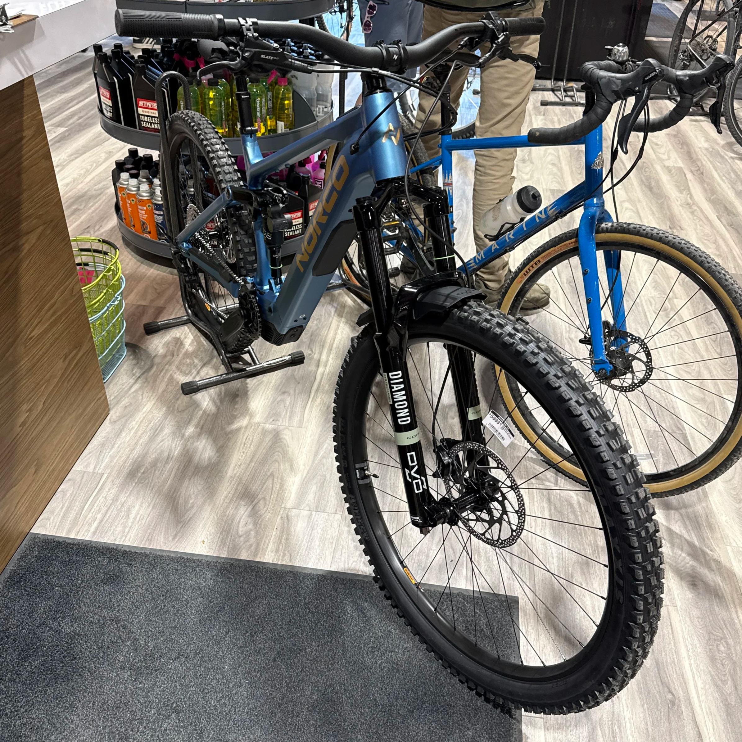 A blue mountain bike inside a cycling shop, with a black wheel featuring thick knobby tires in the foreground and a bike rack in the background filled with yellow spray bottles, bottles of lubricant, and other bike maintenance supplies.