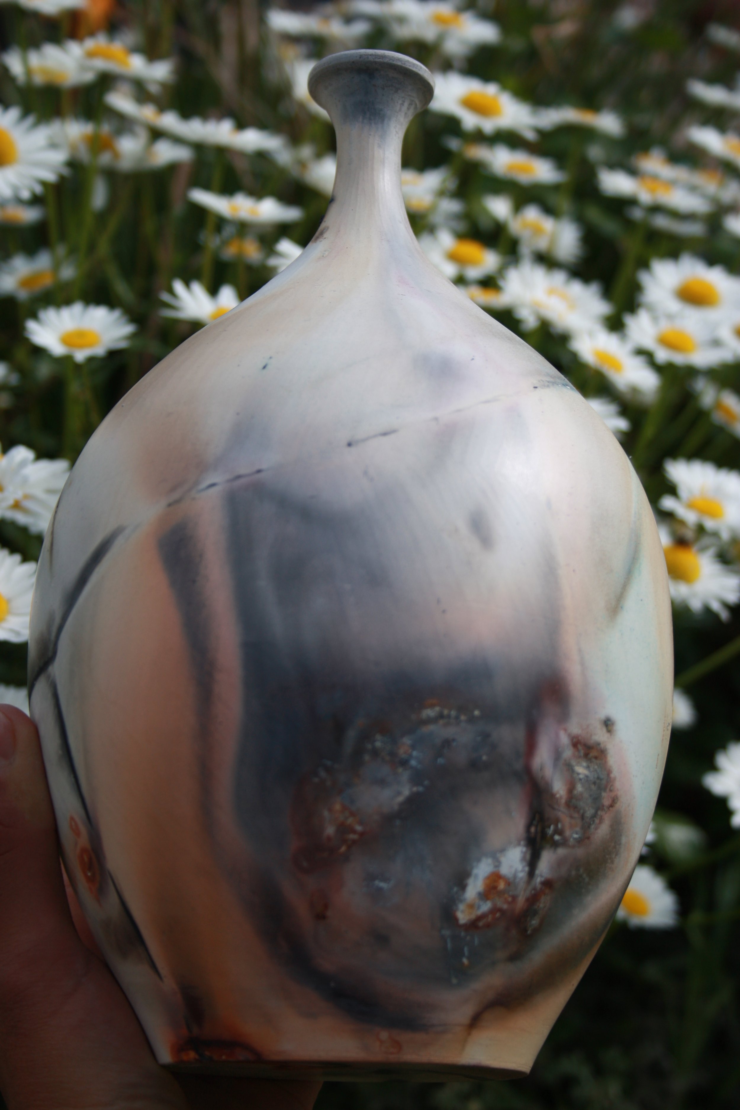 Close-up of a finished pit fired pot with a rounded, bulbous shape and a narrow neck, held by a hand against a background of white daisies with yellow centers.