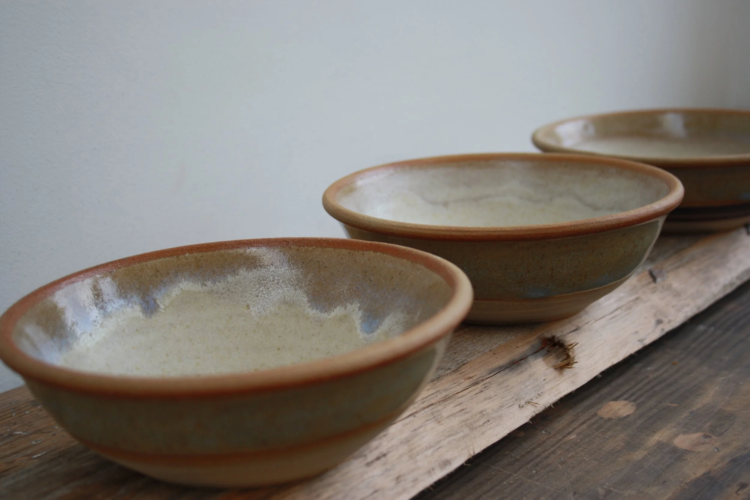 A row of ceramic bowls with a glazed interior and unglazed rim resting on a rustic wooden surface against a plain light background.
