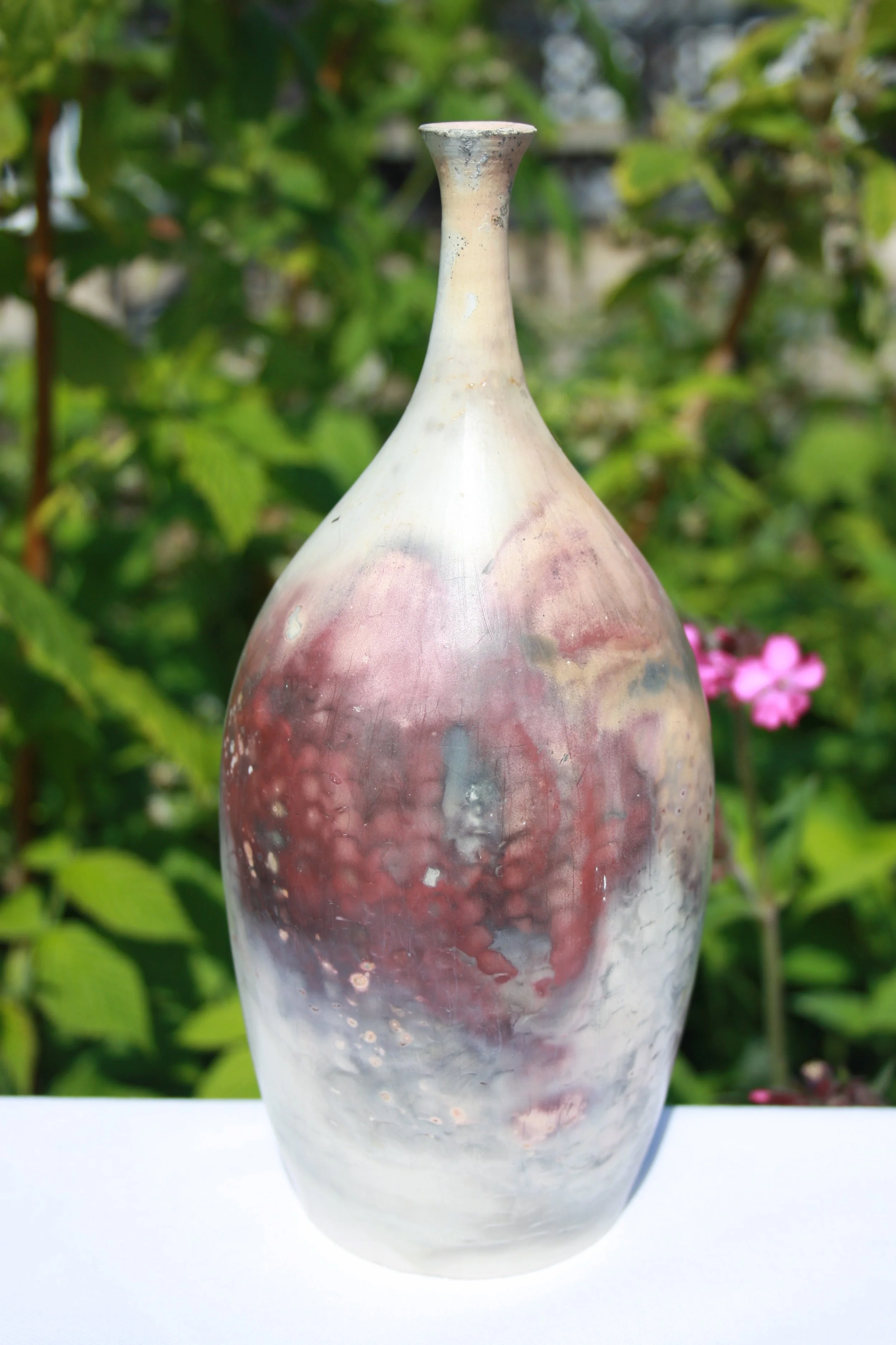 A tall, elongated ceramic vase with a narrow top, featuring a marbled pattern of pink, white, and gray colors, set outdoors with green foliage and pink flowers in the background.