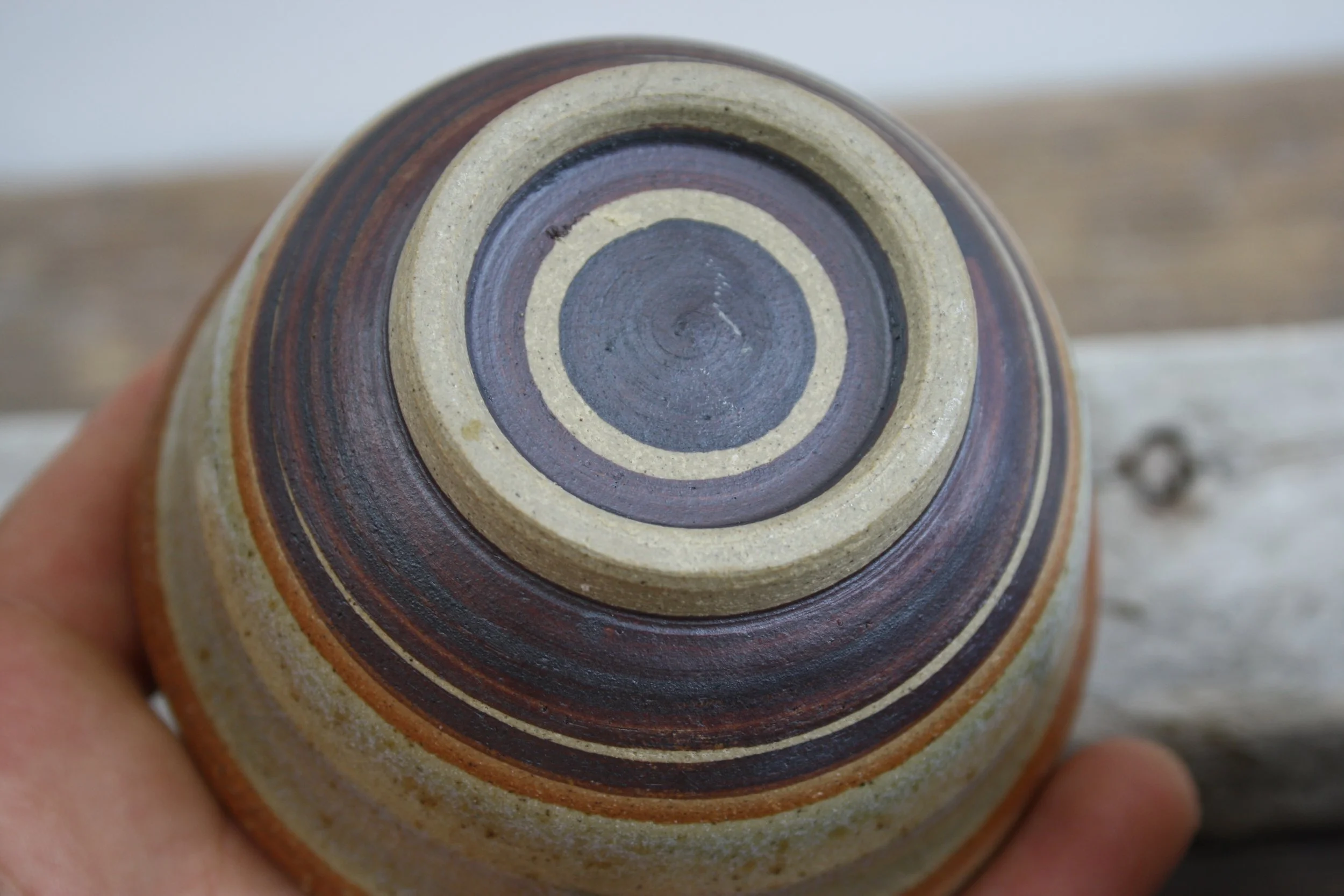 Close-up of the bottom of a ceramic vase or mug with concentric circles in brown, beige, and gray colors, held by a person's hand.